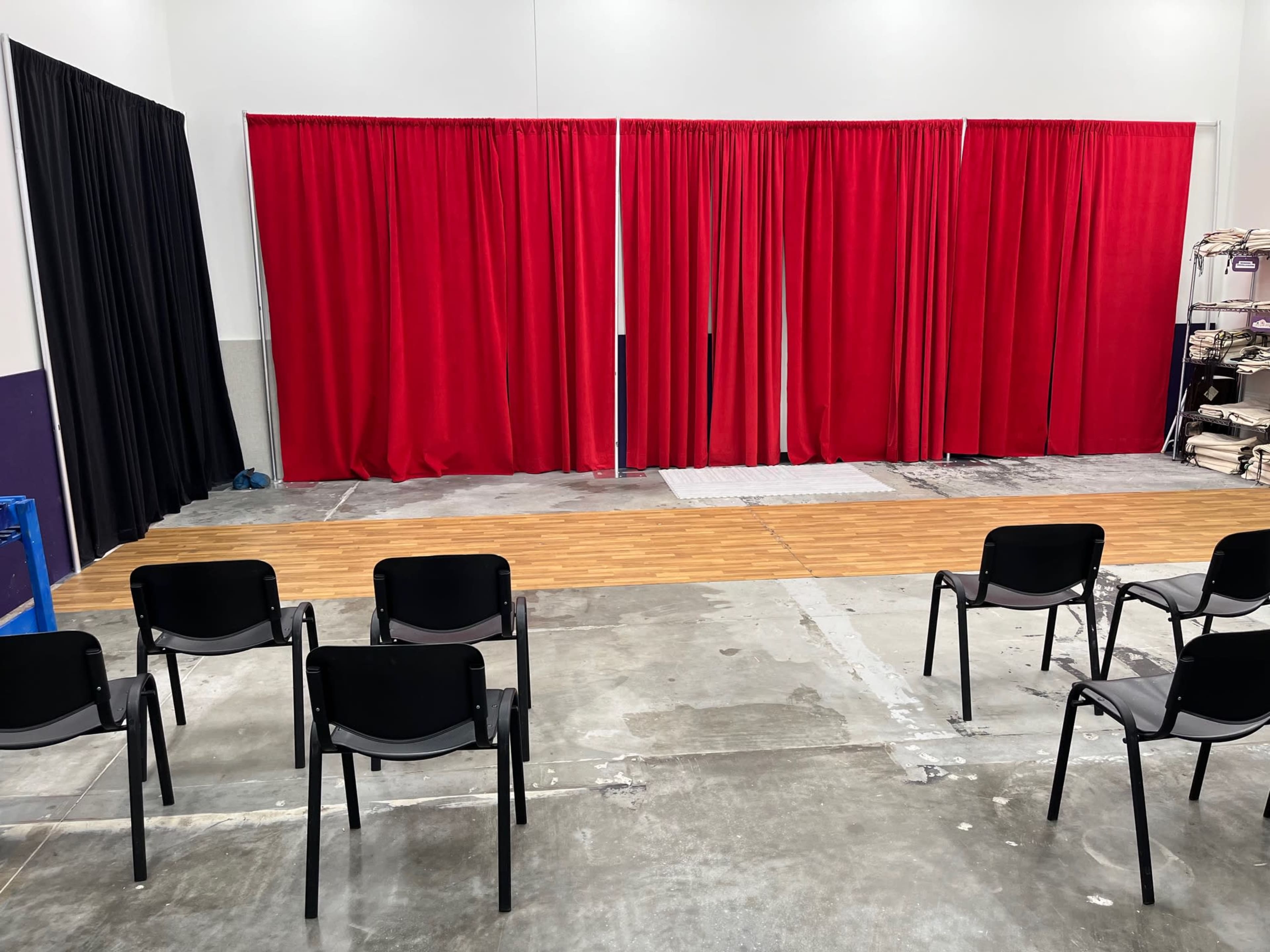 A staged area with red curtains in the center, flanked by black curtains, and a row of black chairs arranged in front on a concrete floor.