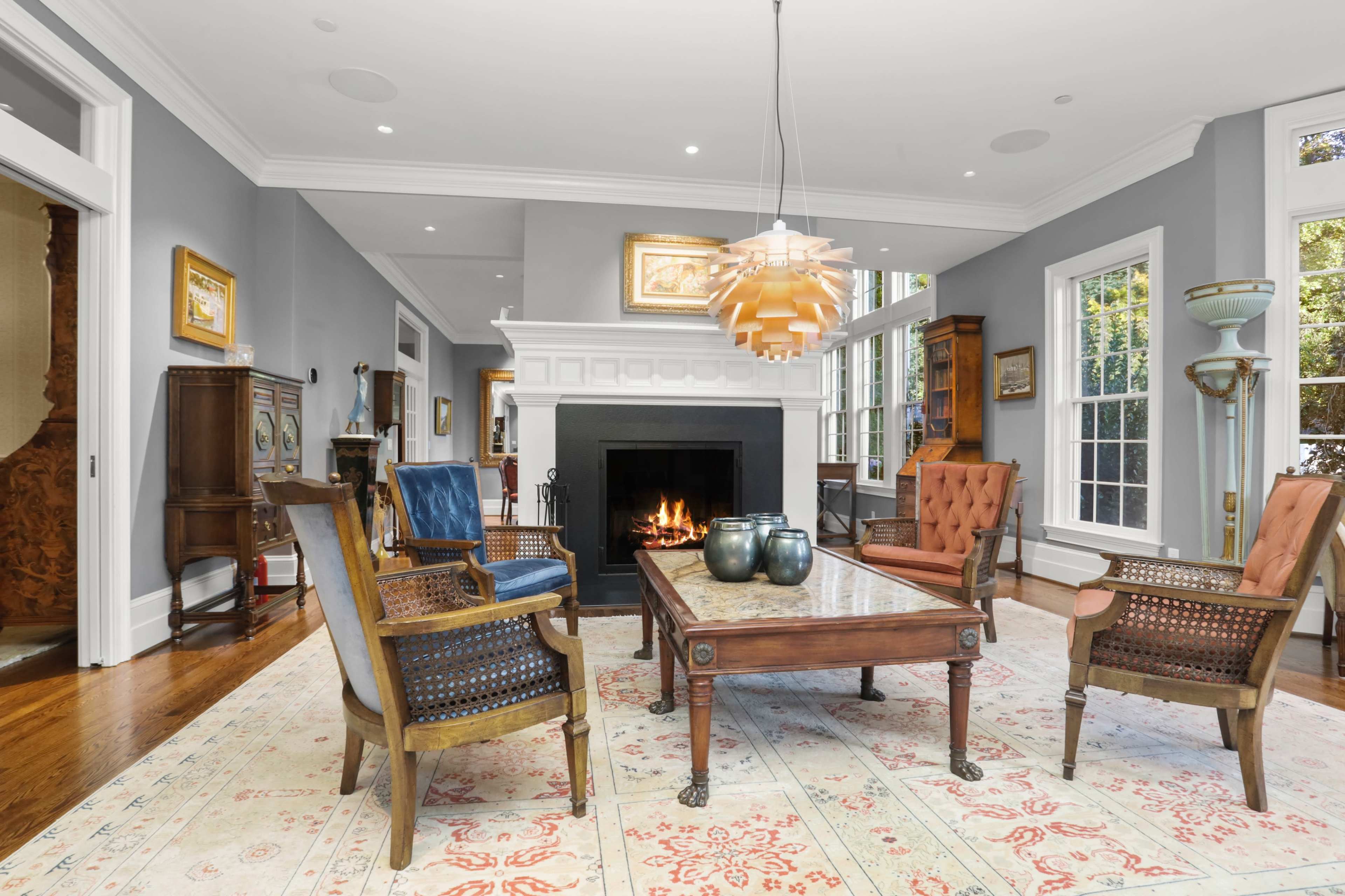 The room features a fireplace surrounded by four chairs and a coffee table on a patterned rug, with large windows allowing natural light.