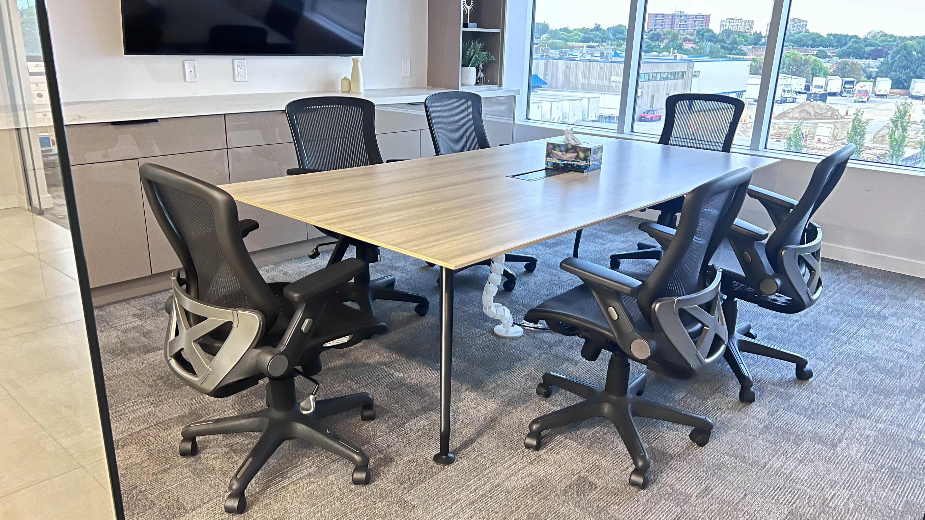 The image shows a modern conference room with a long wooden table surrounded by five ergonomic chairs, and a large screen mounted on the wall.