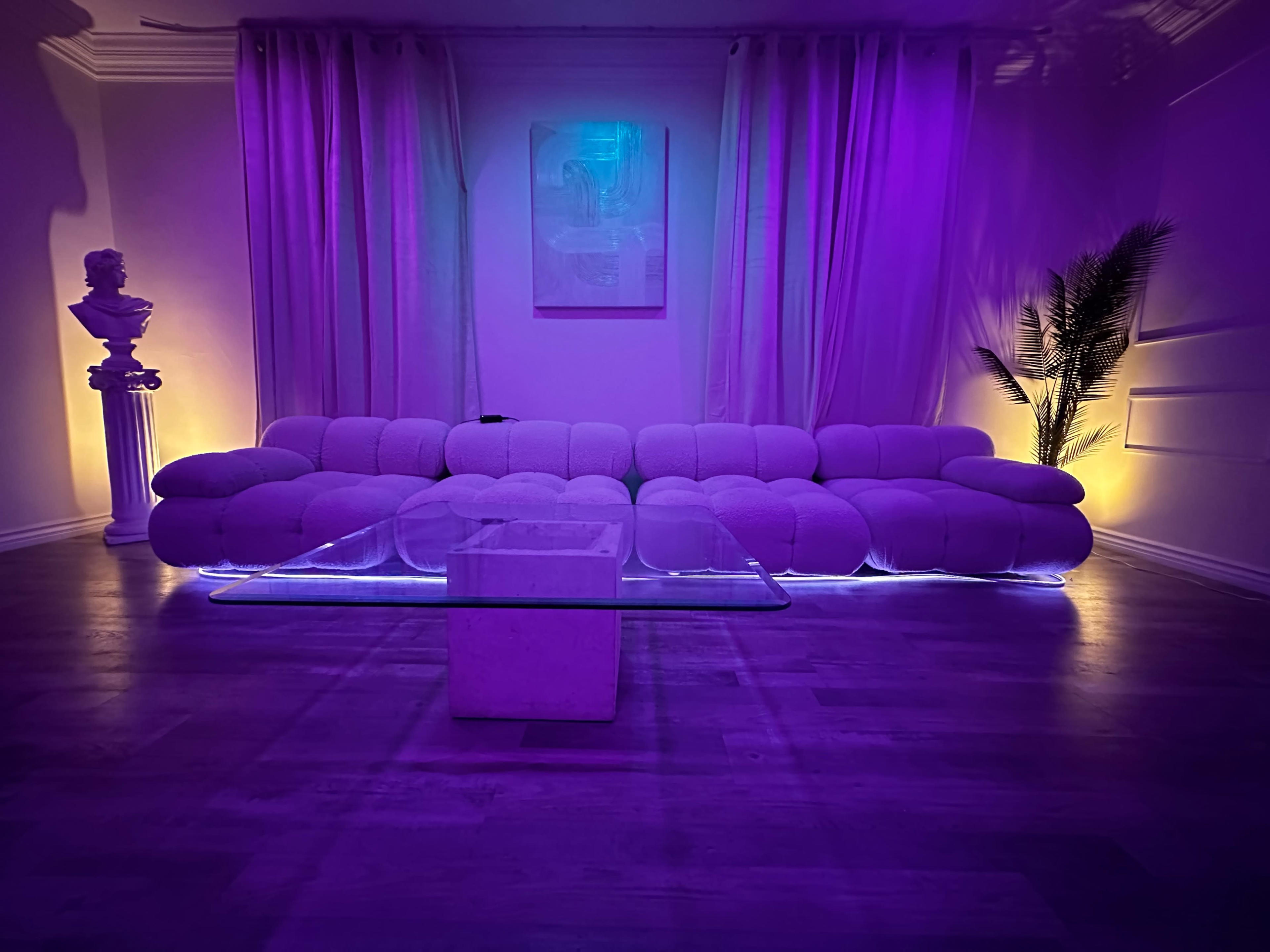 The image shows a modern living room featuring a plush sofa with a glass coffee table, illuminated by purple ambient lighting and complemented by decorative elements like a potted plant and a statue.