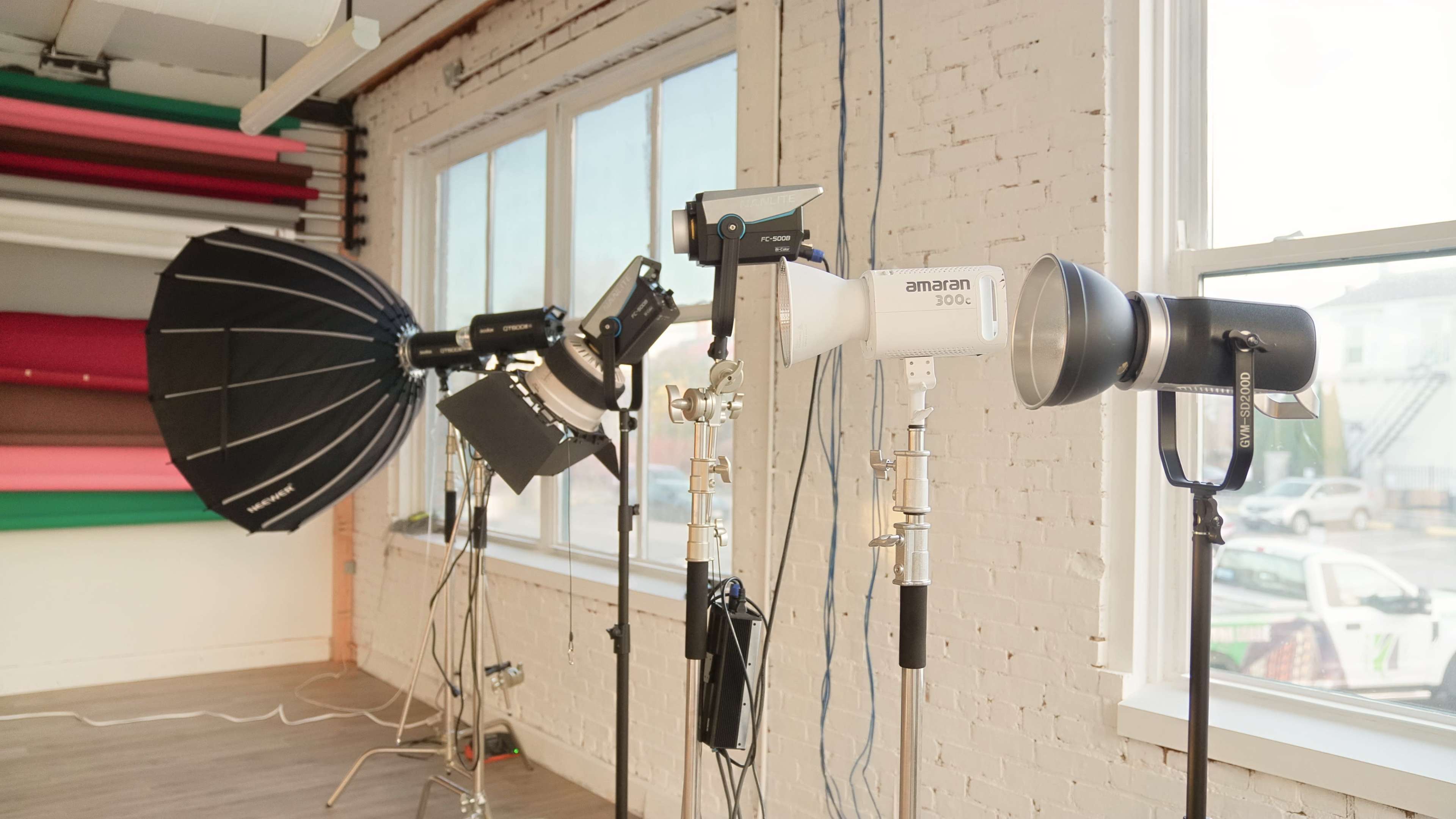 The image shows a photography studio setup with various lighting equipment arranged near large windows, including softboxes and flash units.