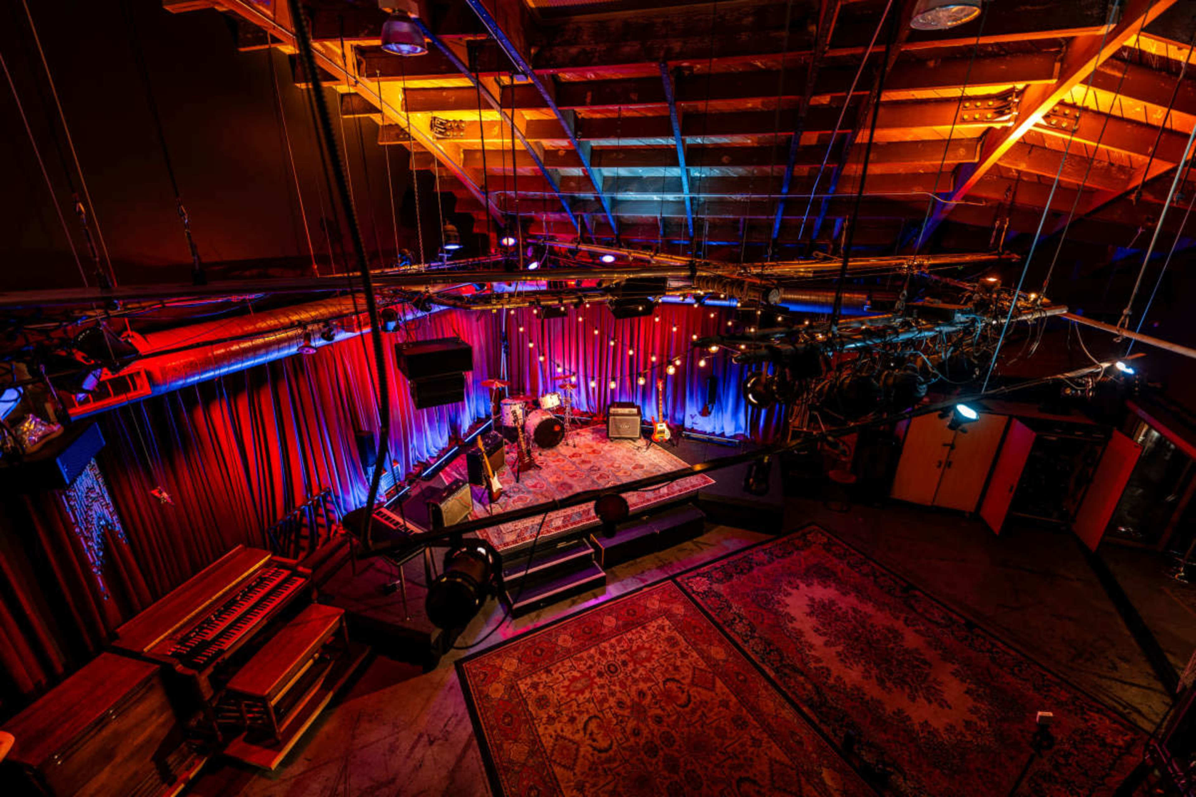 The image shows a dimly lit performance space with vibrant colored lights, a raised stage, and musical equipment arranged on the floor.