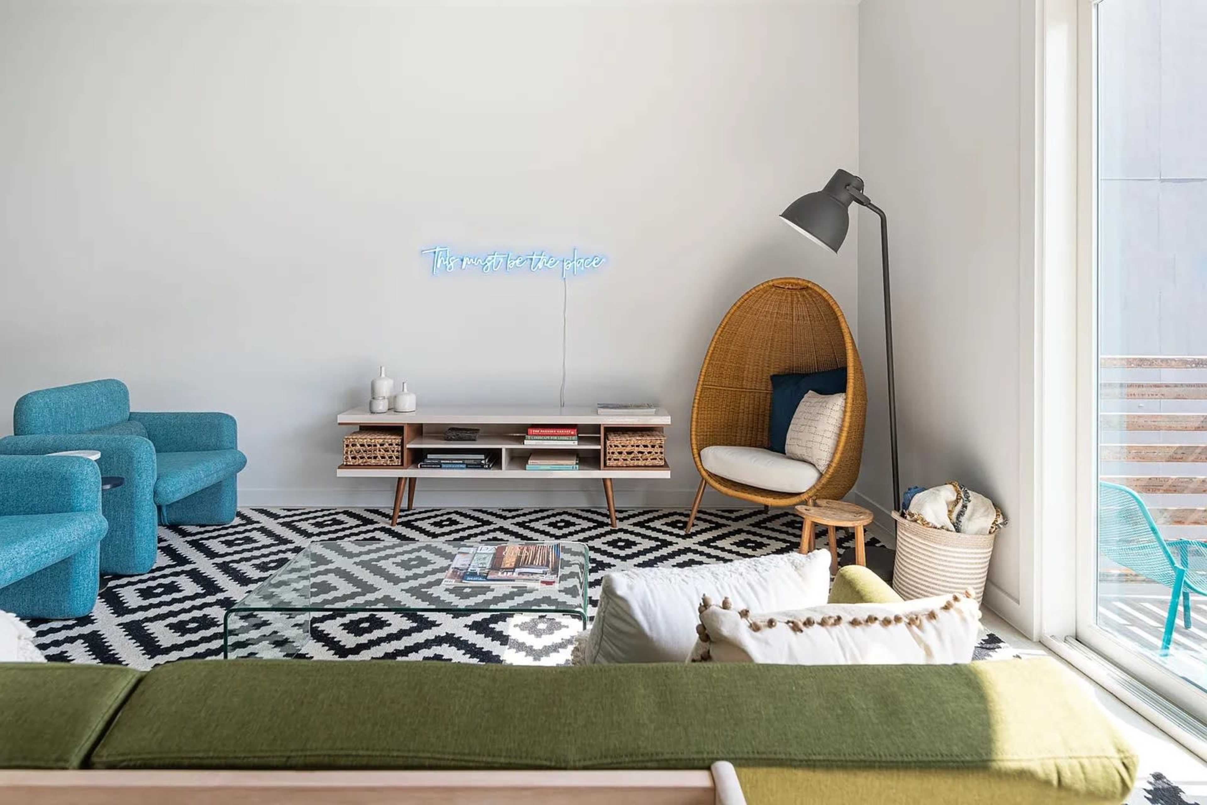 A modern living room with a patterned rug, a green sofa, a blue armchair, a rattan chair, and a minimalist coffee table.