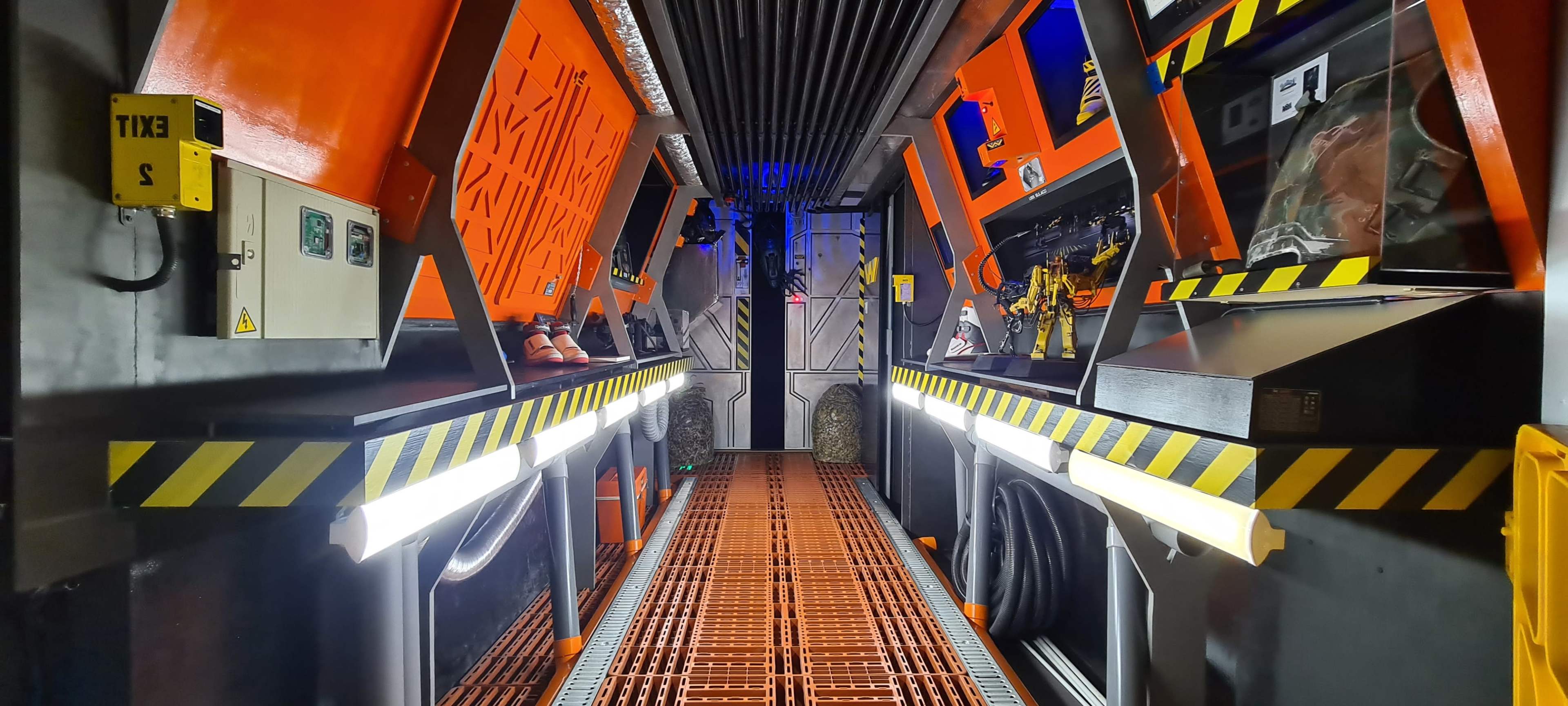 A futuristic interior corridor with orange and black walls, illuminated by fluorescent lights and lined with various equipment and gear on the sides.