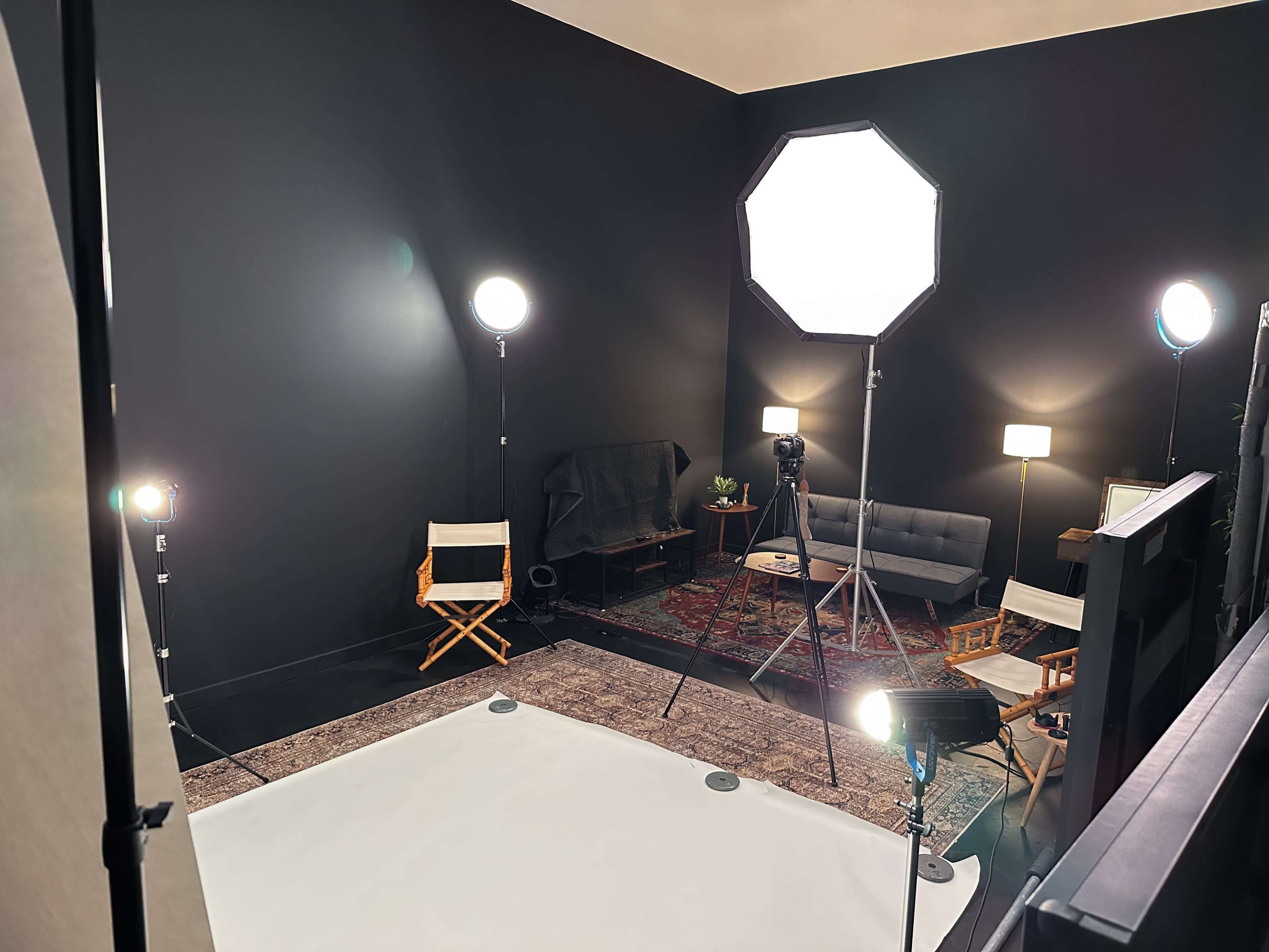 The image shows a photography studio with multiple lighting setups, a gray sofa, and a large softbox light.