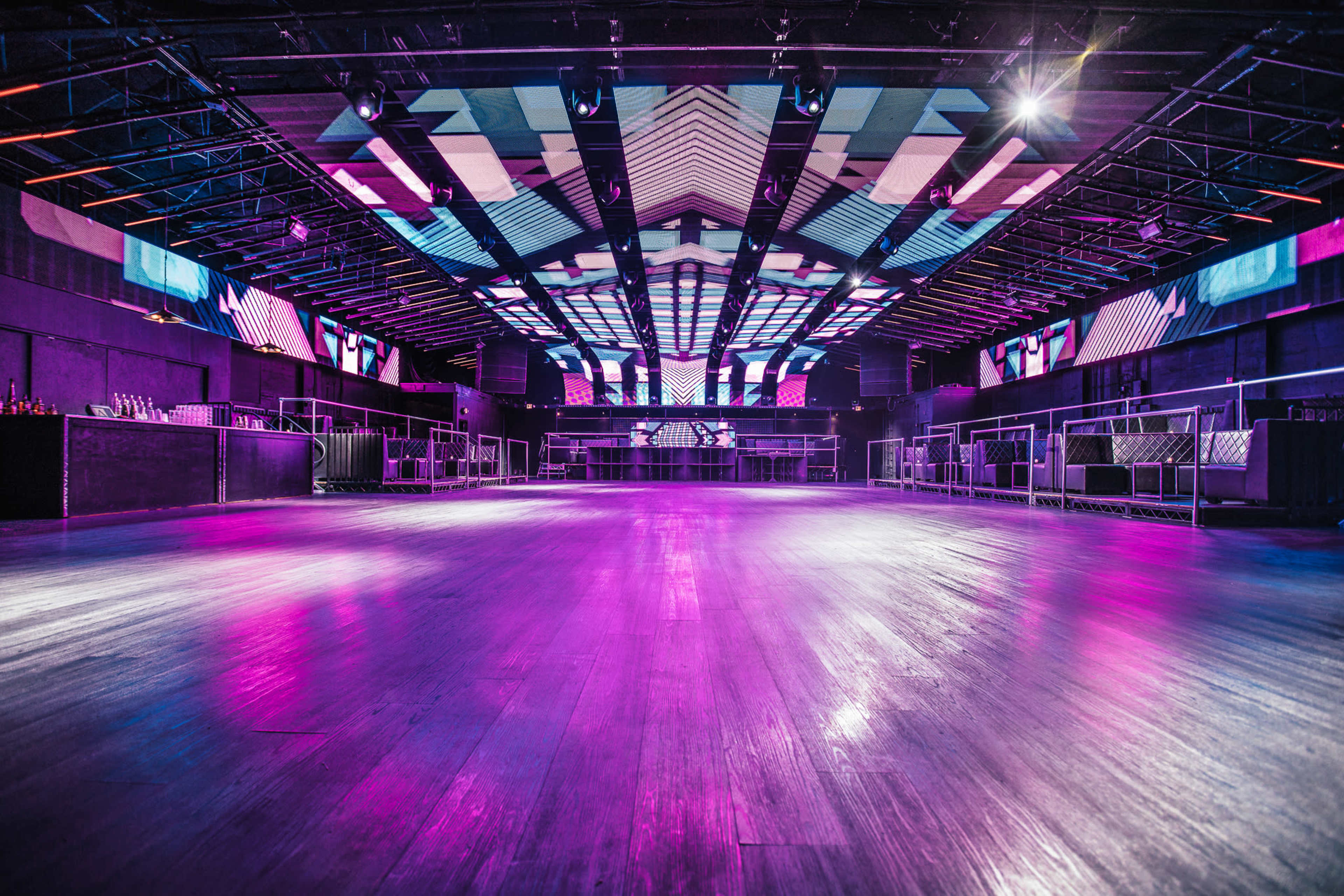 The image shows an empty nightclub interior with a polished wooden floor, colorful LED screens on the walls, and a DJ booth at the back.