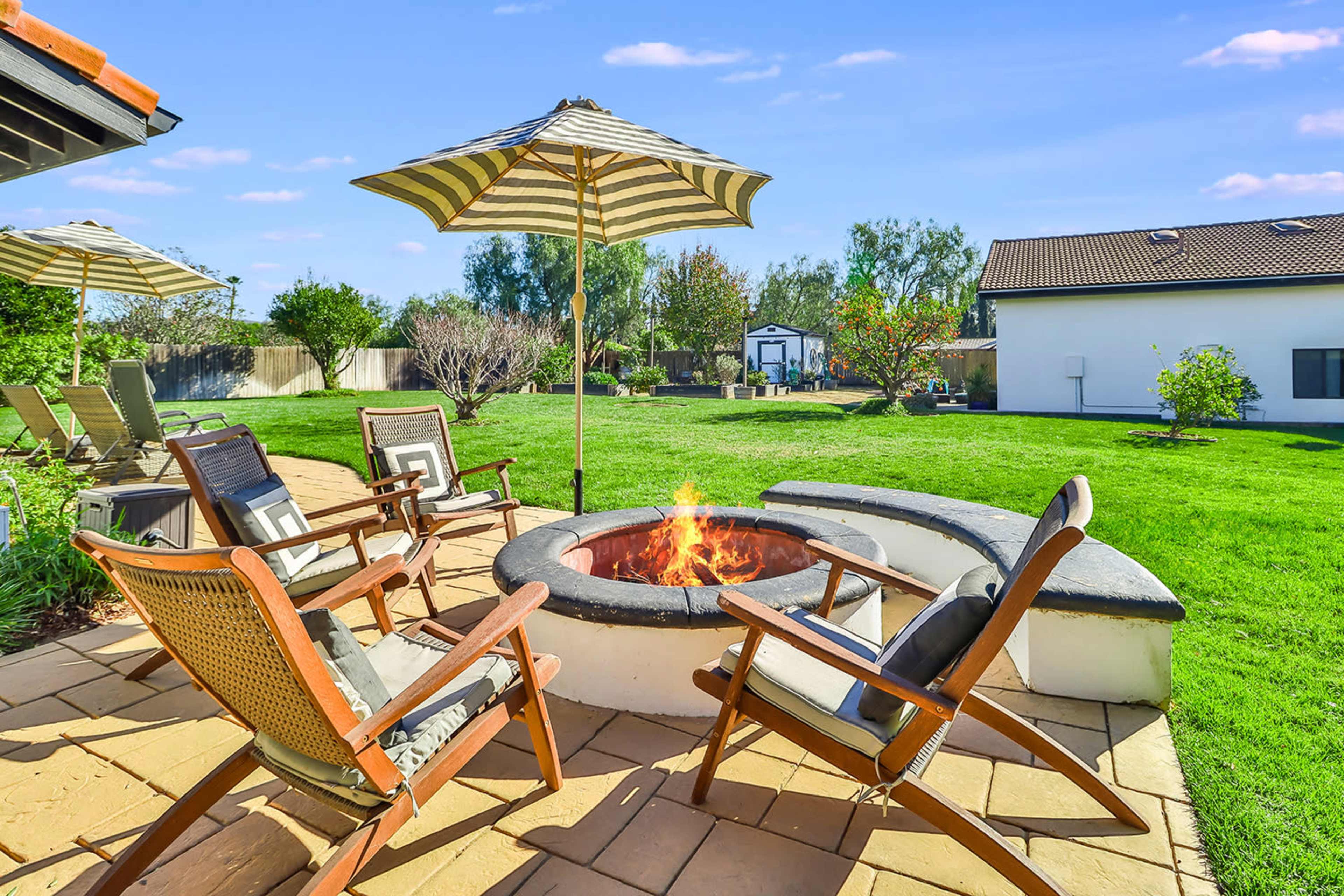 A circular fire pit surrounded by wooden lounge chairs and a striped umbrella is set on a grassy backyard.