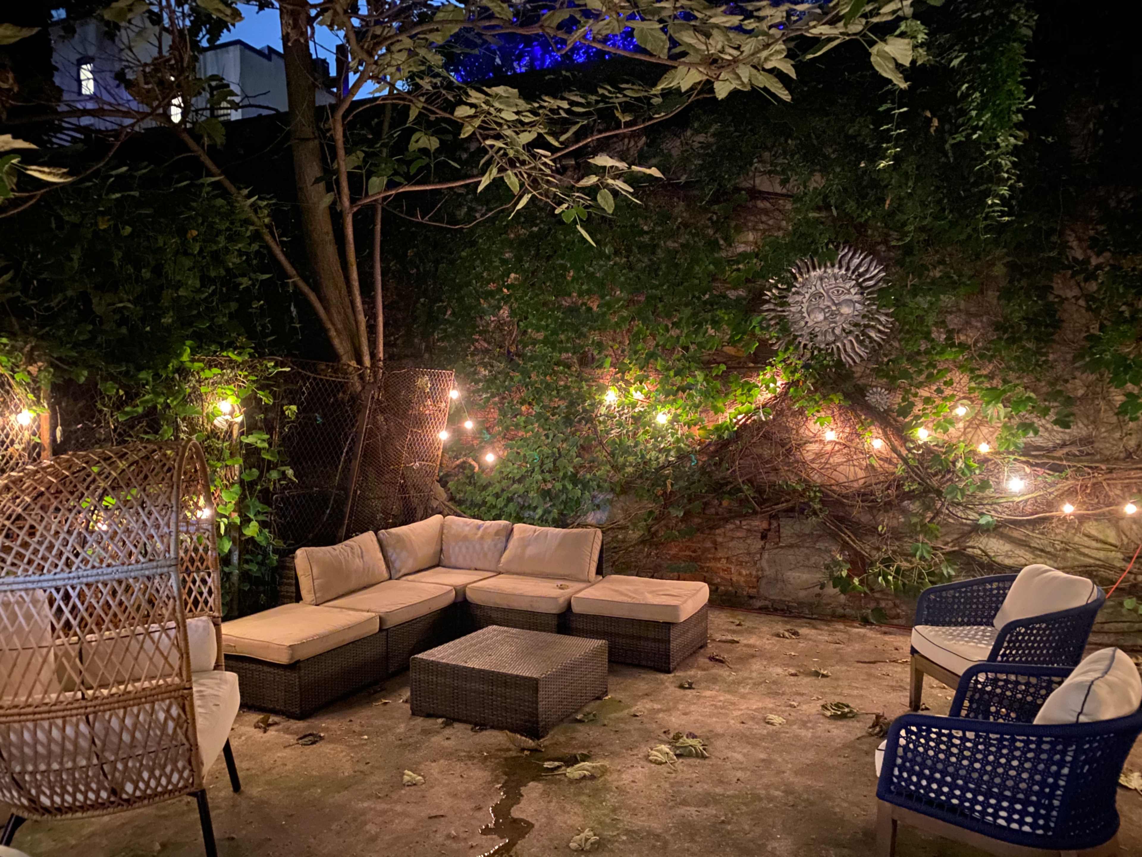 The image shows a garden patio area with a sectional sofa, coffee table, and several chairs surrounded by greenery and string lights.