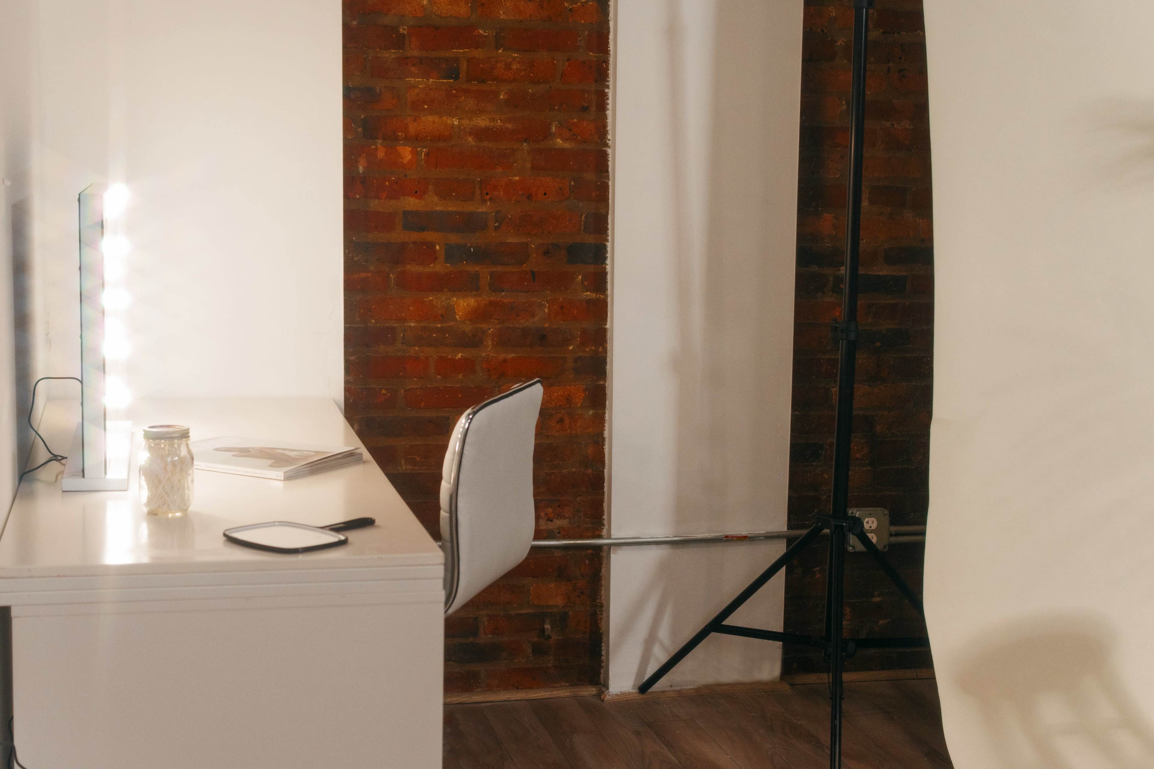 A well-lit workspace features a white desk with a mirror and a chair against a brick wall, accompanied by a backdrop stand and soft lighting.