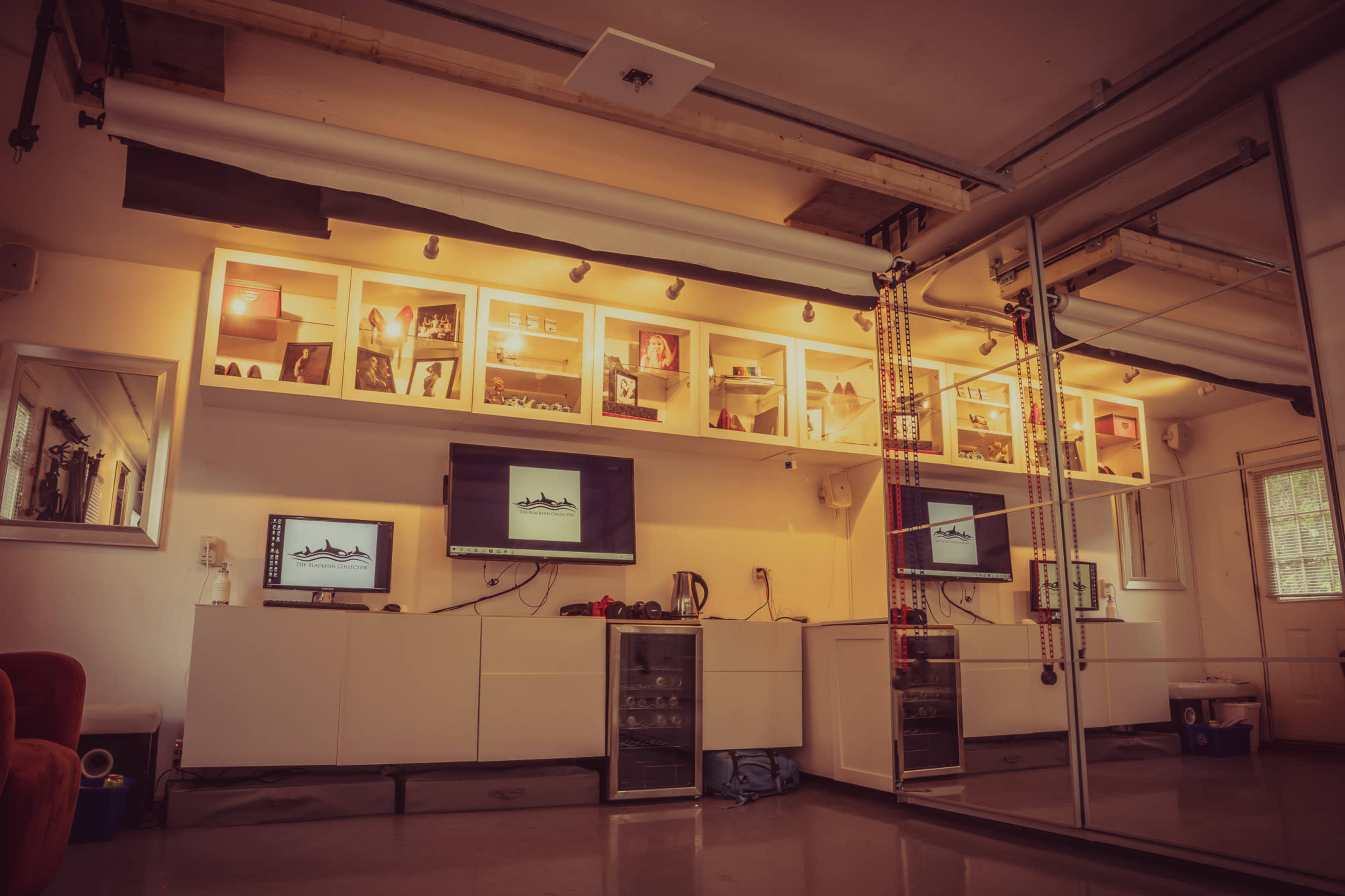 A modern room with a wall-mounted shelving unit, two flat-screen monitors, and a small refrigerator beneath them.