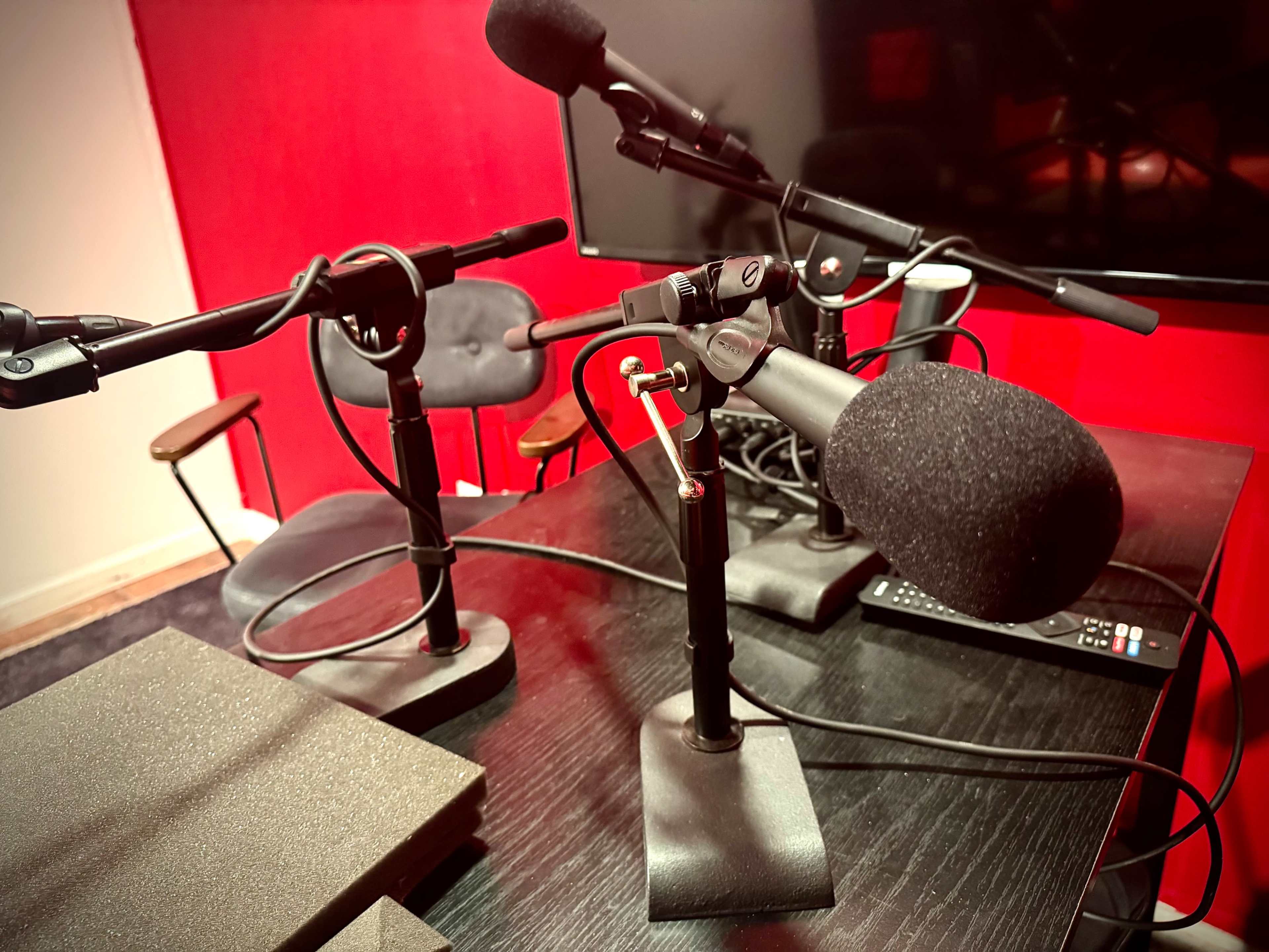 The image shows a podcasting setup with multiple microphones on stands, a black table, and a red wall in the background.