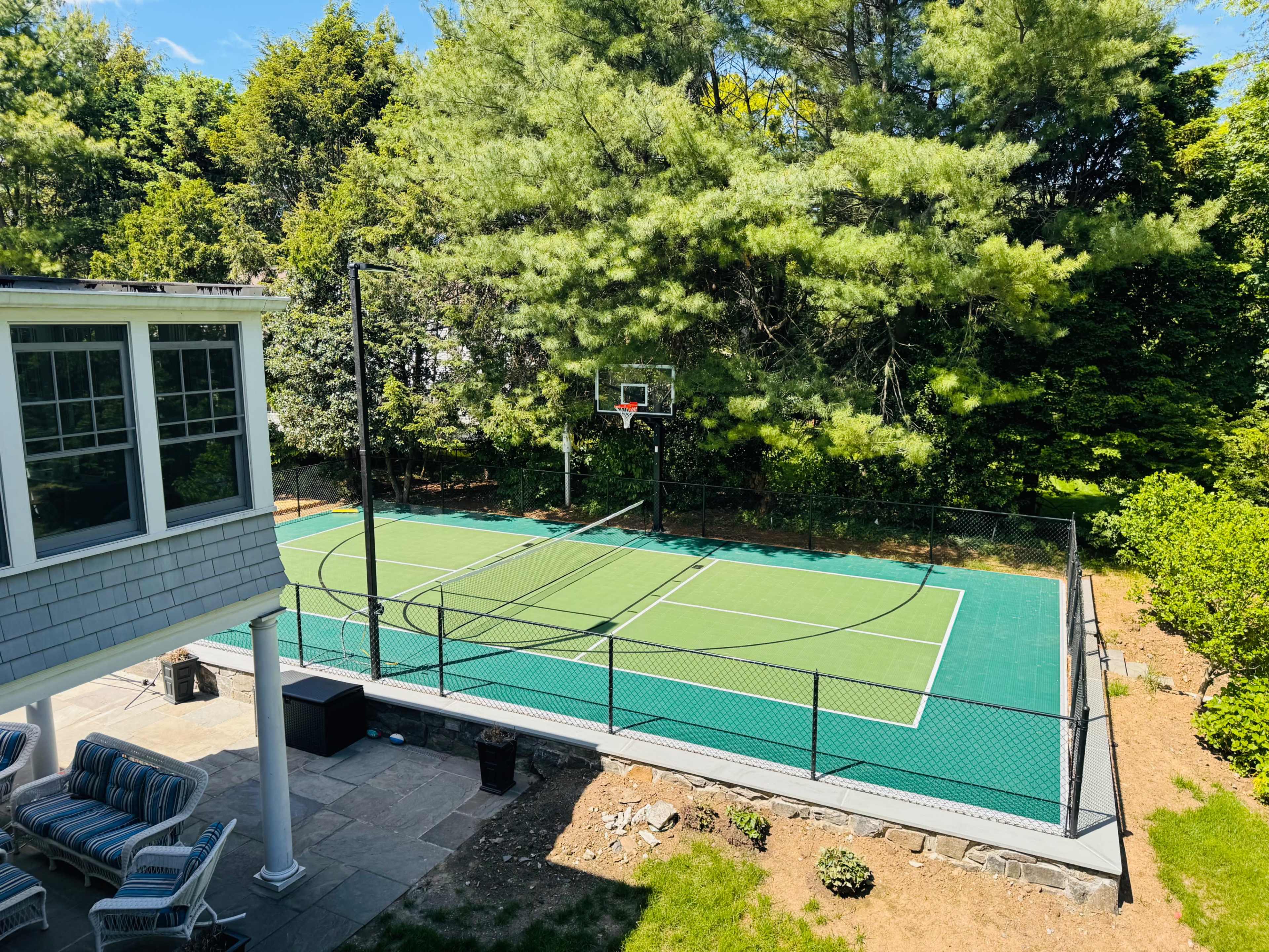 The image shows a well-maintained outdoor basketball court surrounded by trees and a patio area with seating.