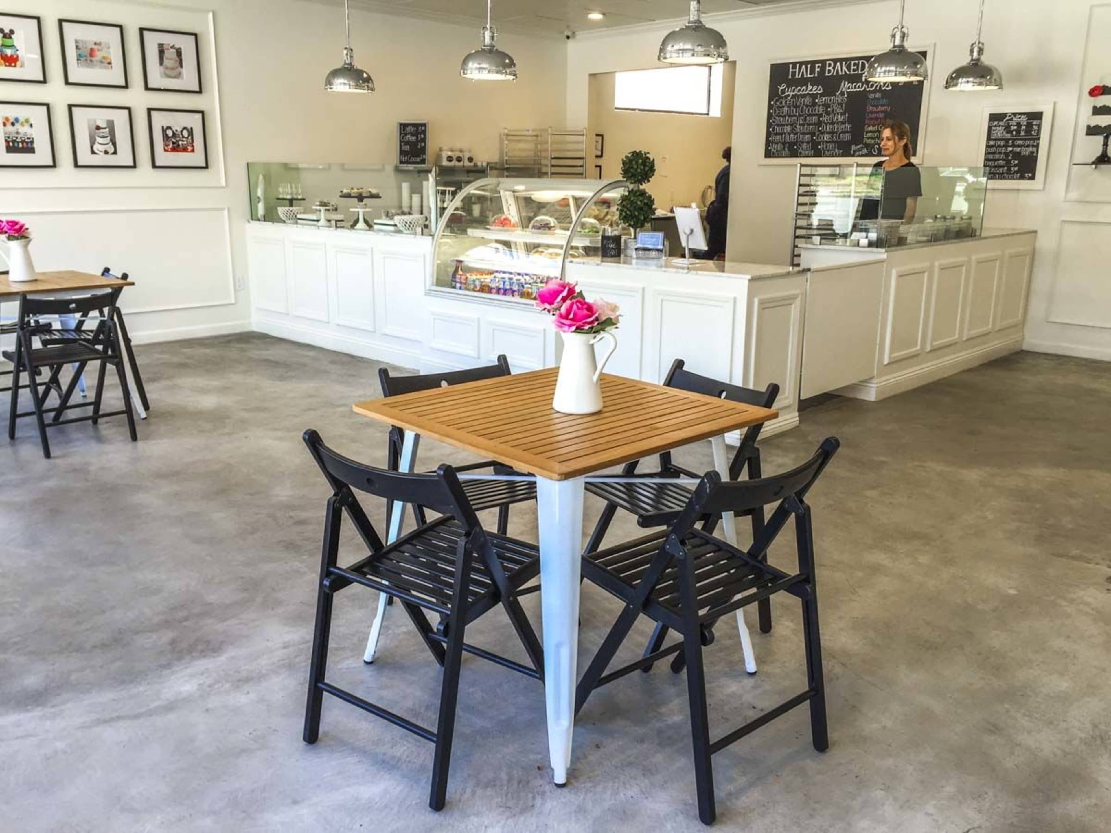 A bright and modern cafe with a wooden table and black folding chairs arranged in the center, complemented by a glass display case of desserts in the background.