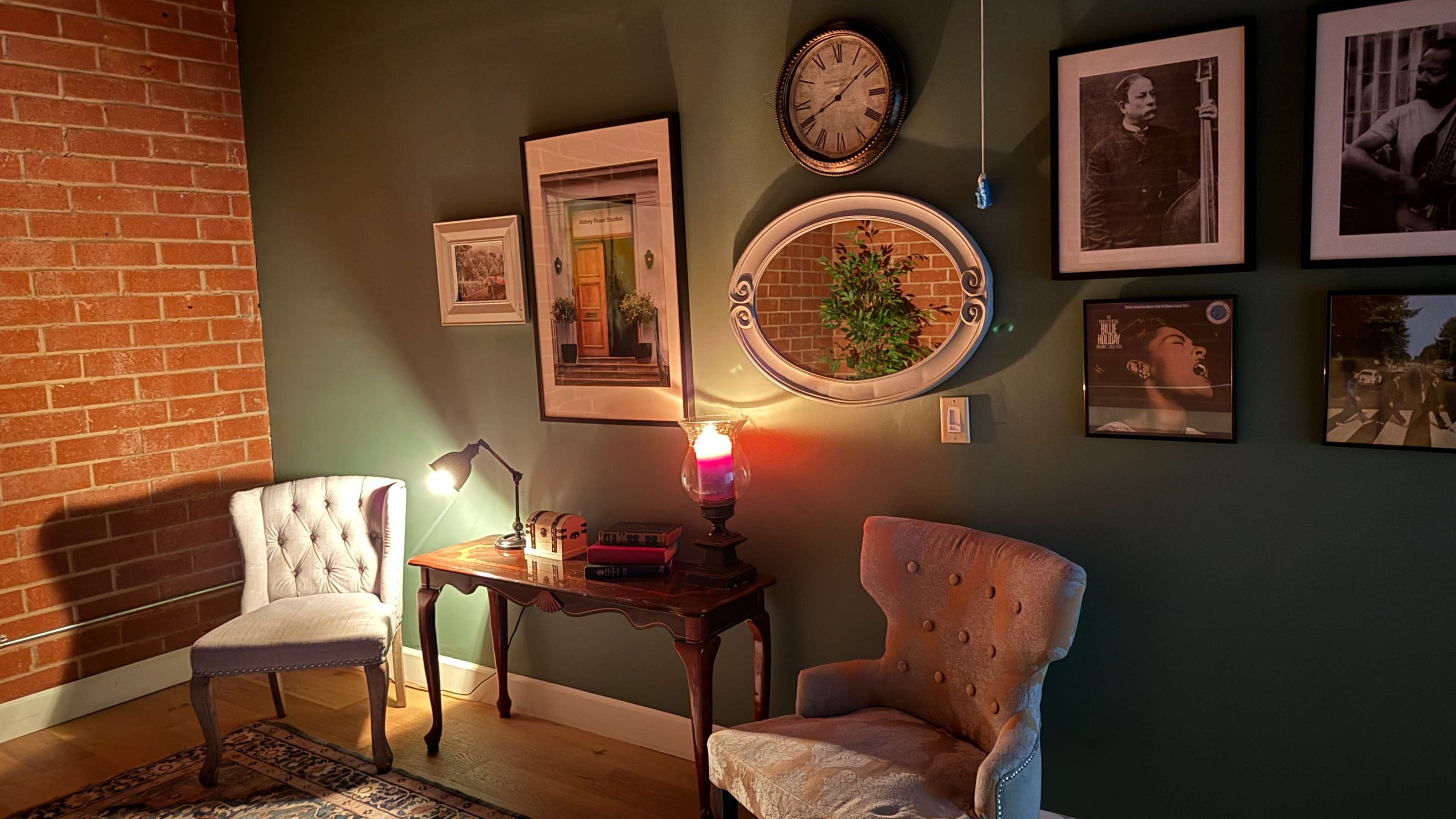The image showcases a cozy corner of a room featuring two upholstered chairs, a small table with a lamp, and a collection of framed photos on a green wall.