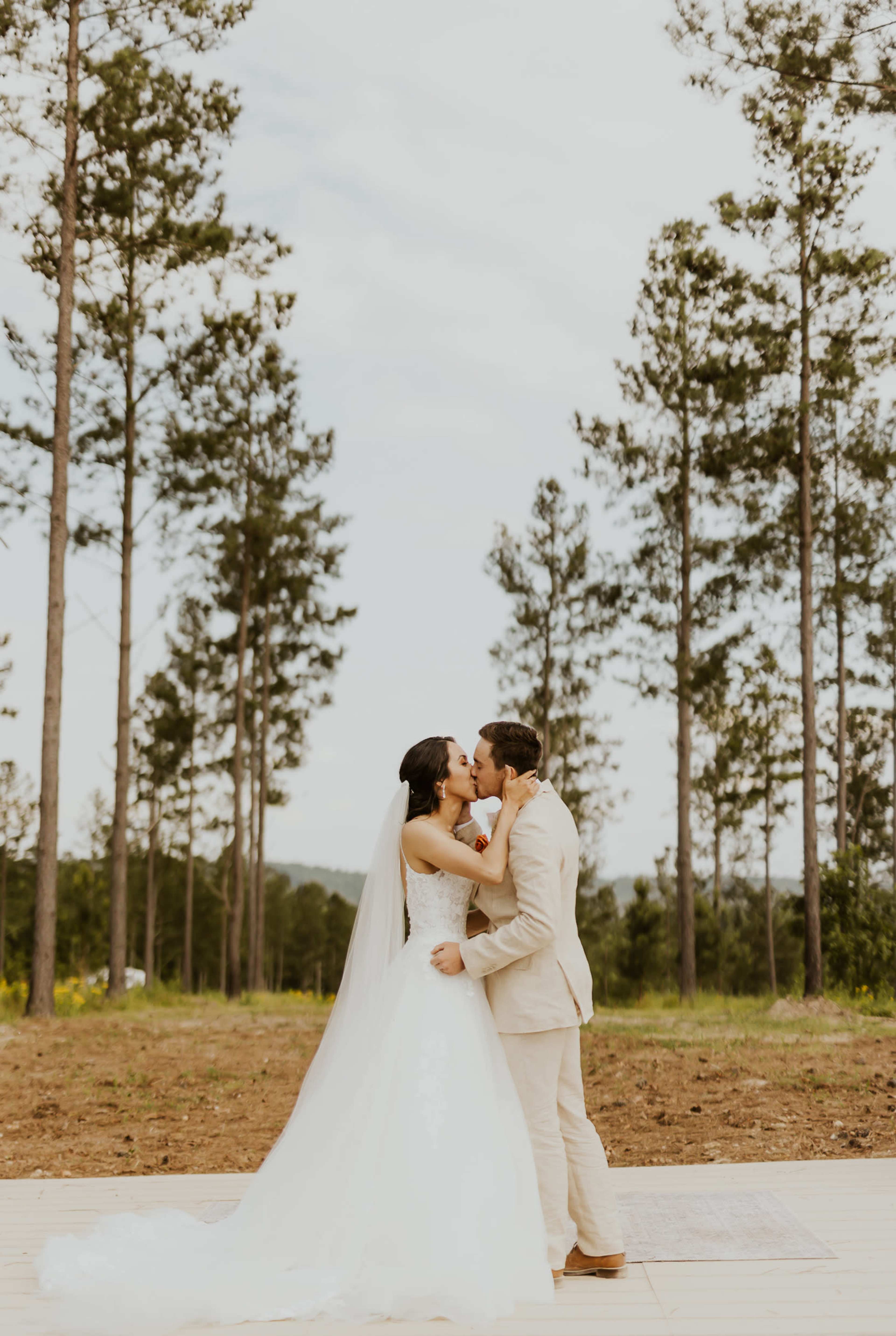 A bride and groom kiss in a forest clearing surrounded by tall pine trees.