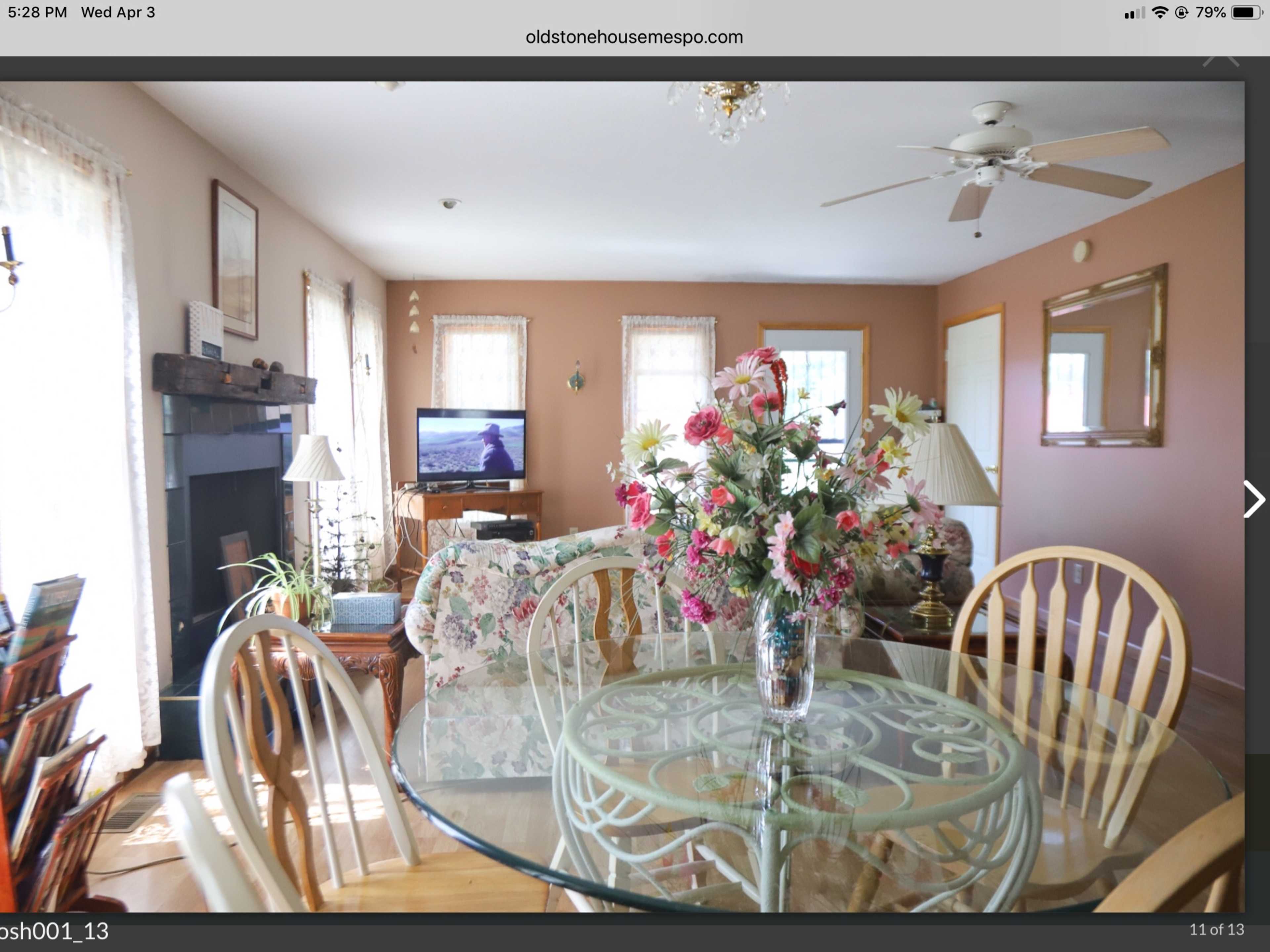 A living room with a glass dining table, floral centerpiece, two chairs, a floral couch, and a television against the wall, with large windows allowing natural light.
