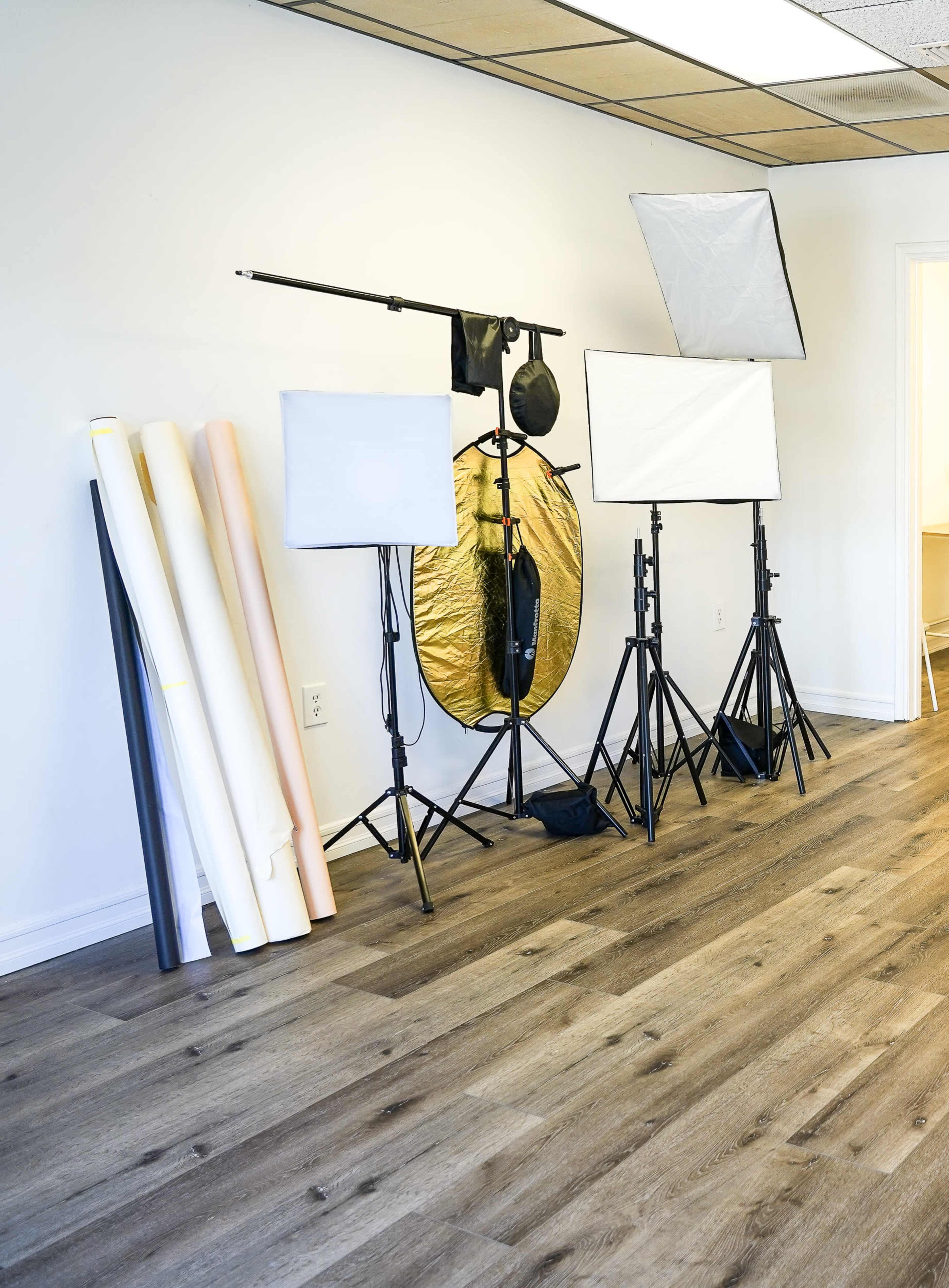 A photography studio setup includes lighting equipment, reflectors, and rolls of backdrop paper against a plain wall.
