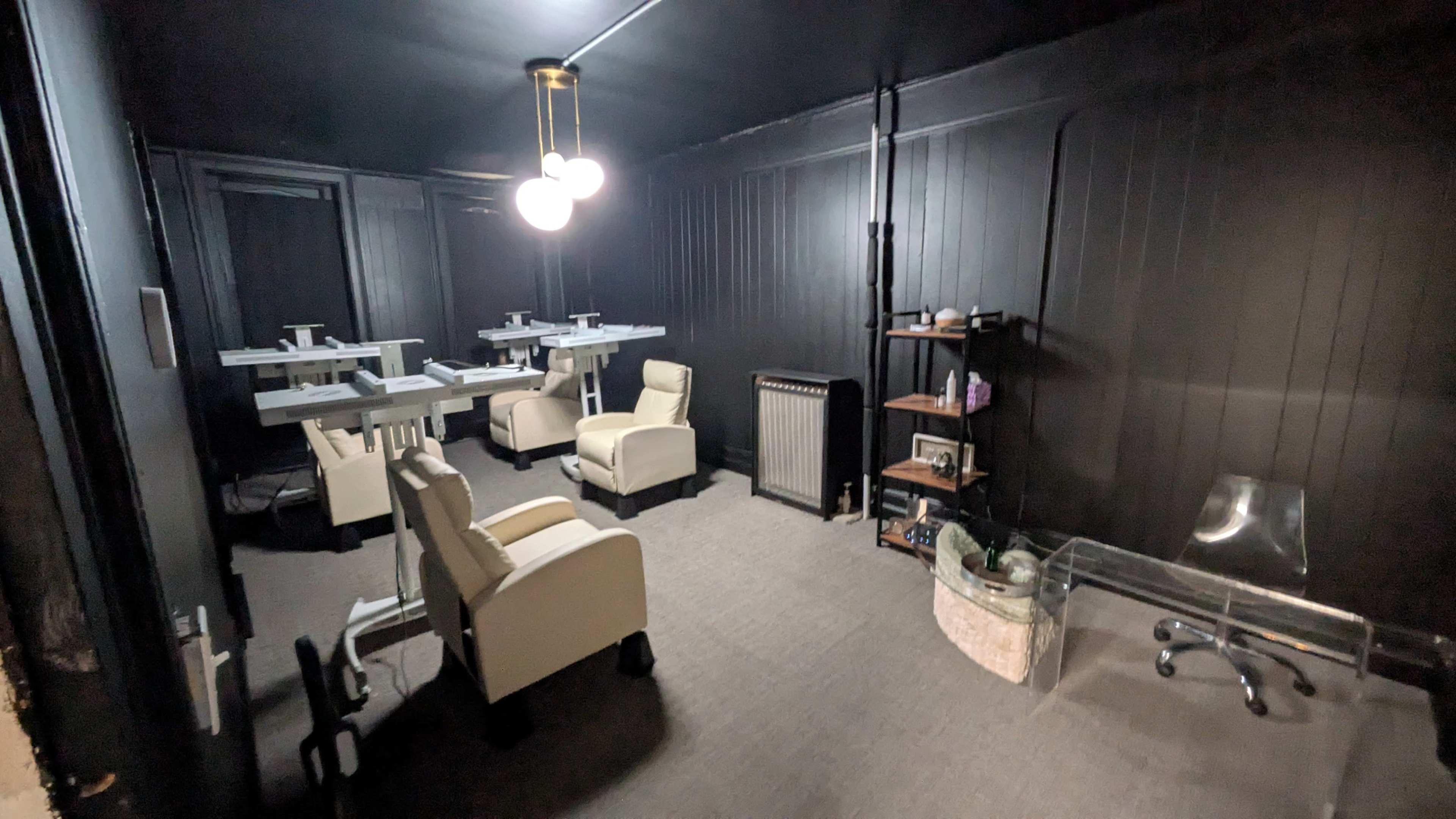 The image shows a dimly lit room with black walls, featuring two white chairs, a glass desk, and a few tables arranged for work or meetings.