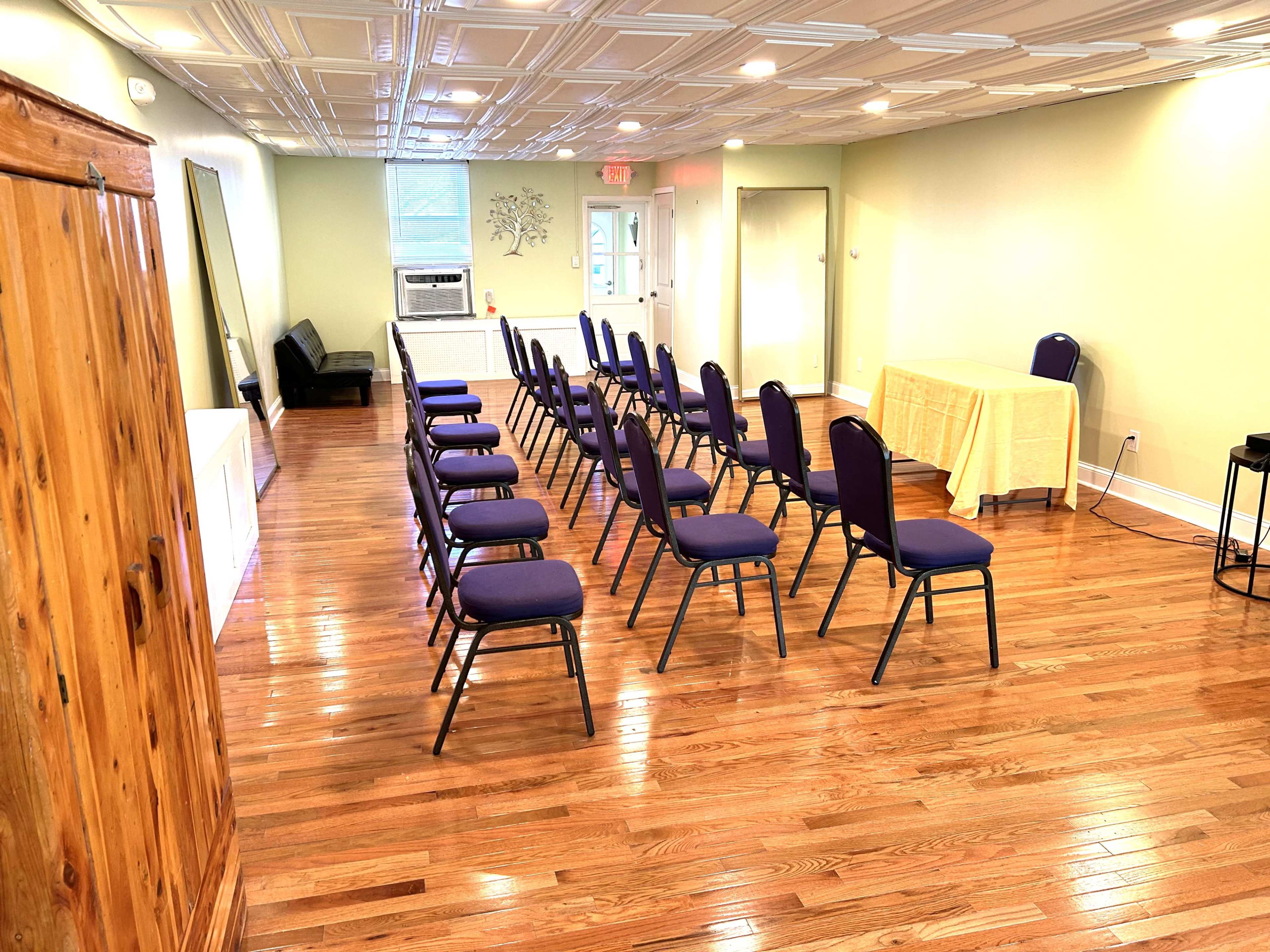 A well-lit room with polished wooden floors, featuring several rows of chairs facing a table in the front.