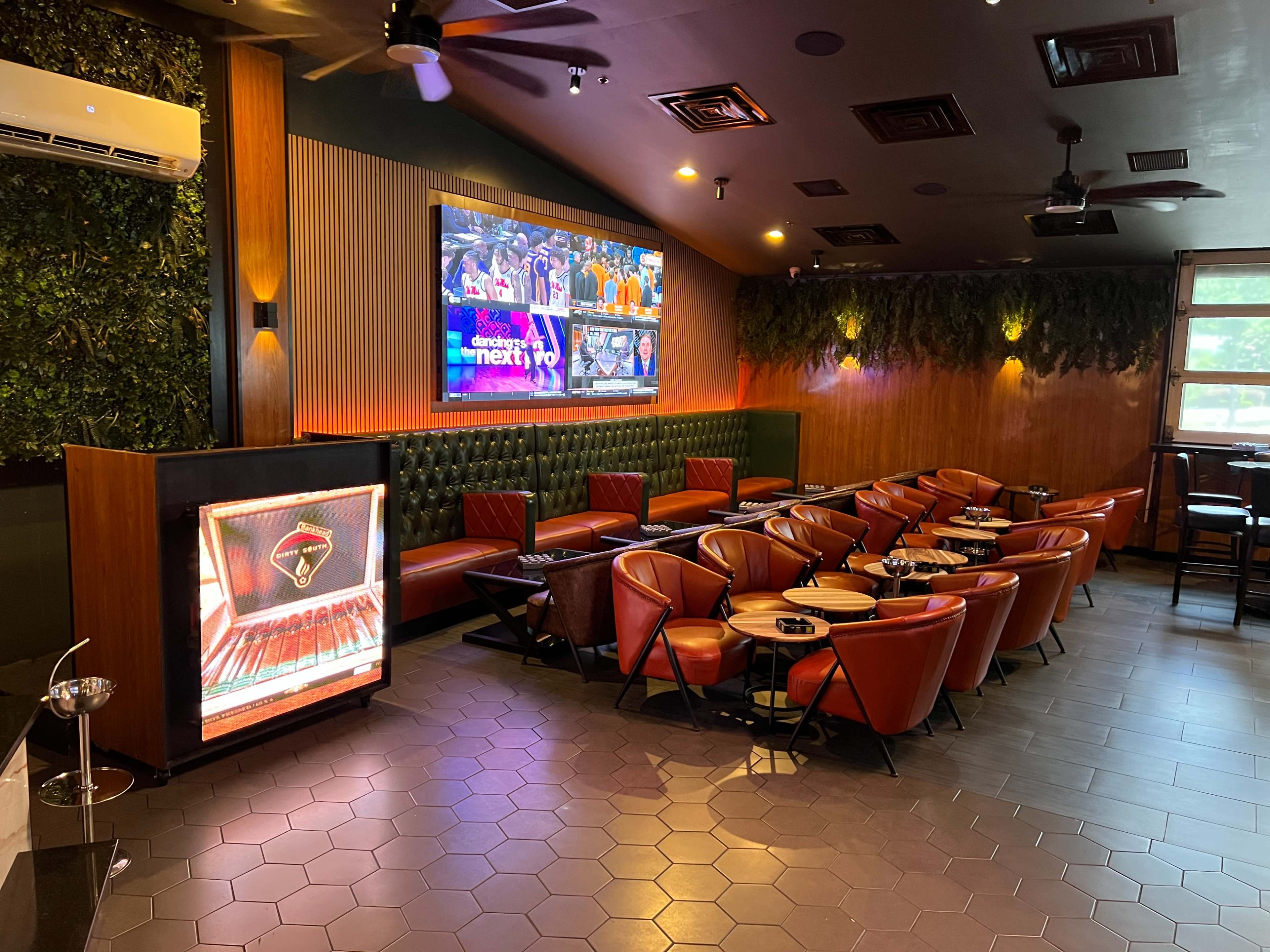 The image shows a modern lounge area with green plants on the wall, red leather chairs arranged around small tables, and a large screen displaying content.