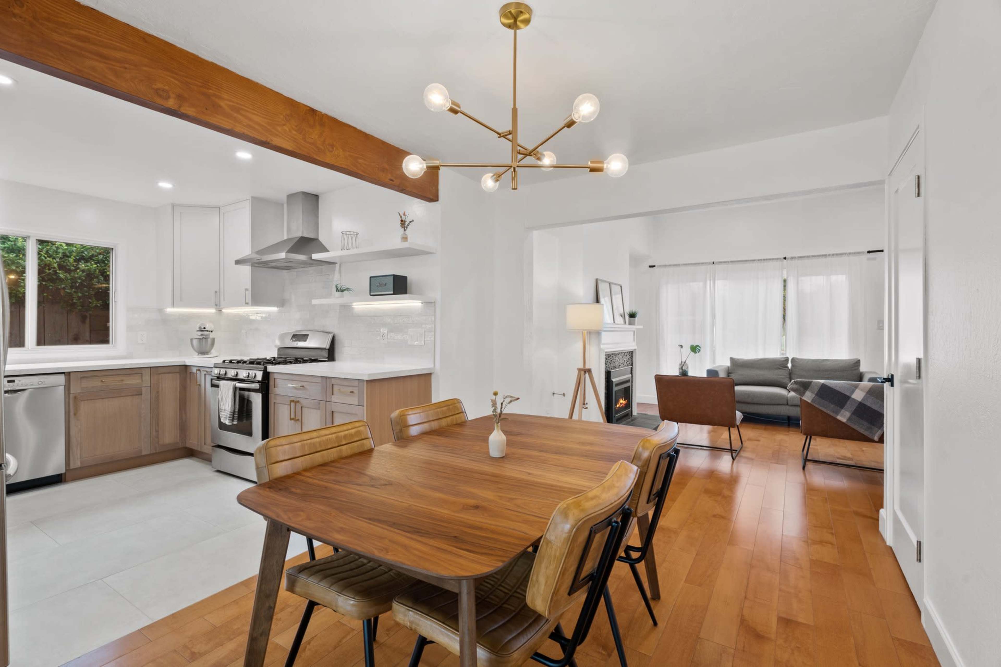 A modern kitchen and dining area featuring a wooden dining table, sleek appliances, and a living room visible in the background with a couch and large windows.