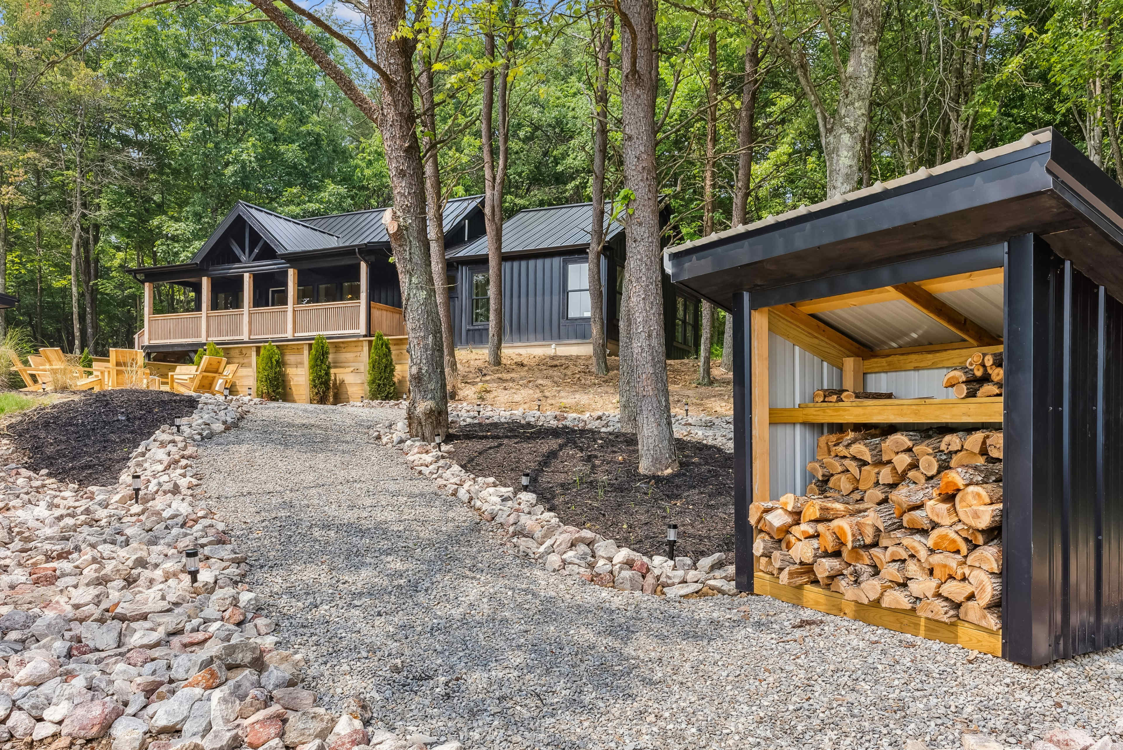 A gravel path leads to a modern cabin surrounded by trees, with a wood storage shed nearby.