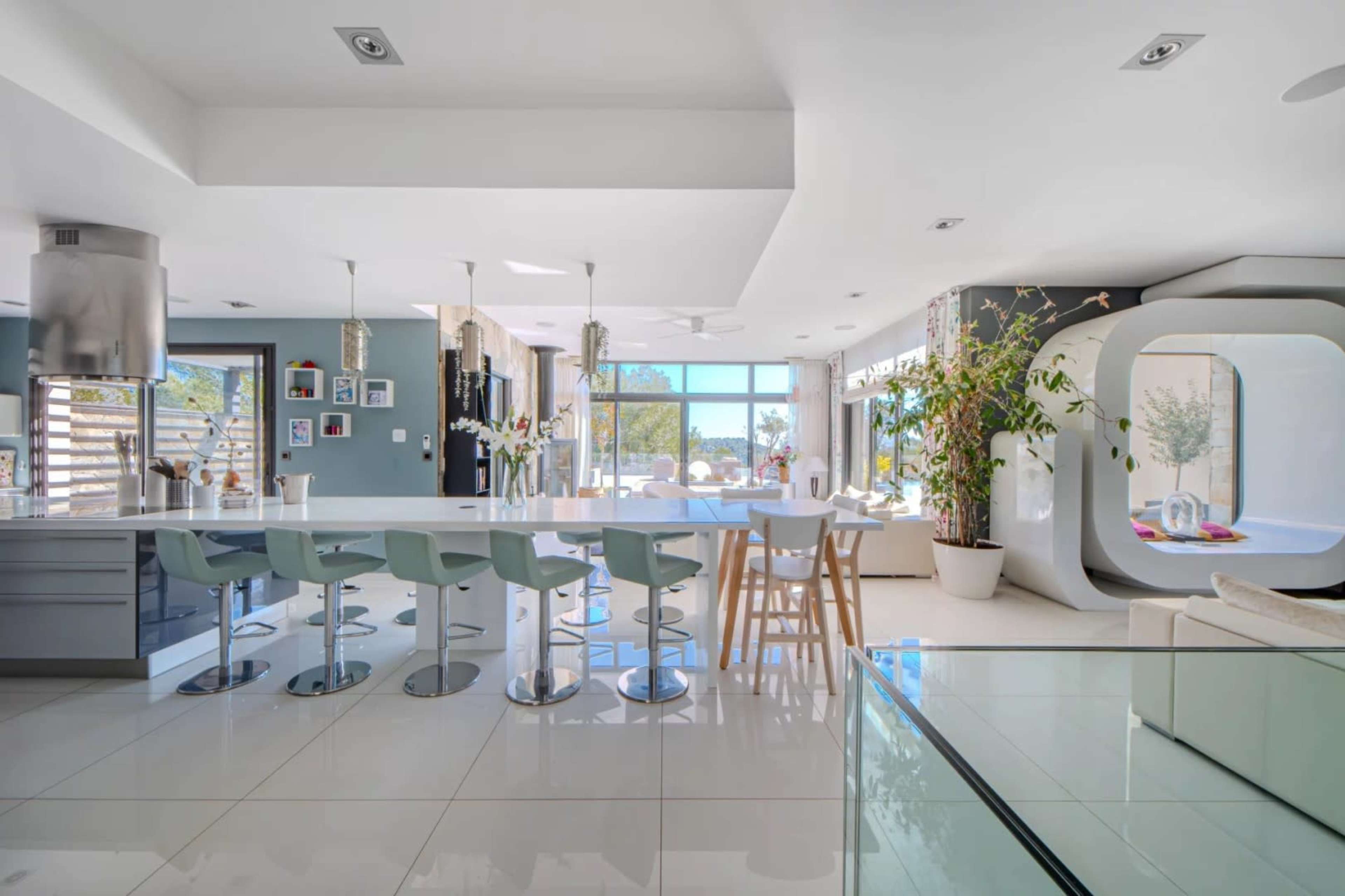 The image shows a modern kitchen and dining area with a large island, bar stools, and abundant natural light from large windows.