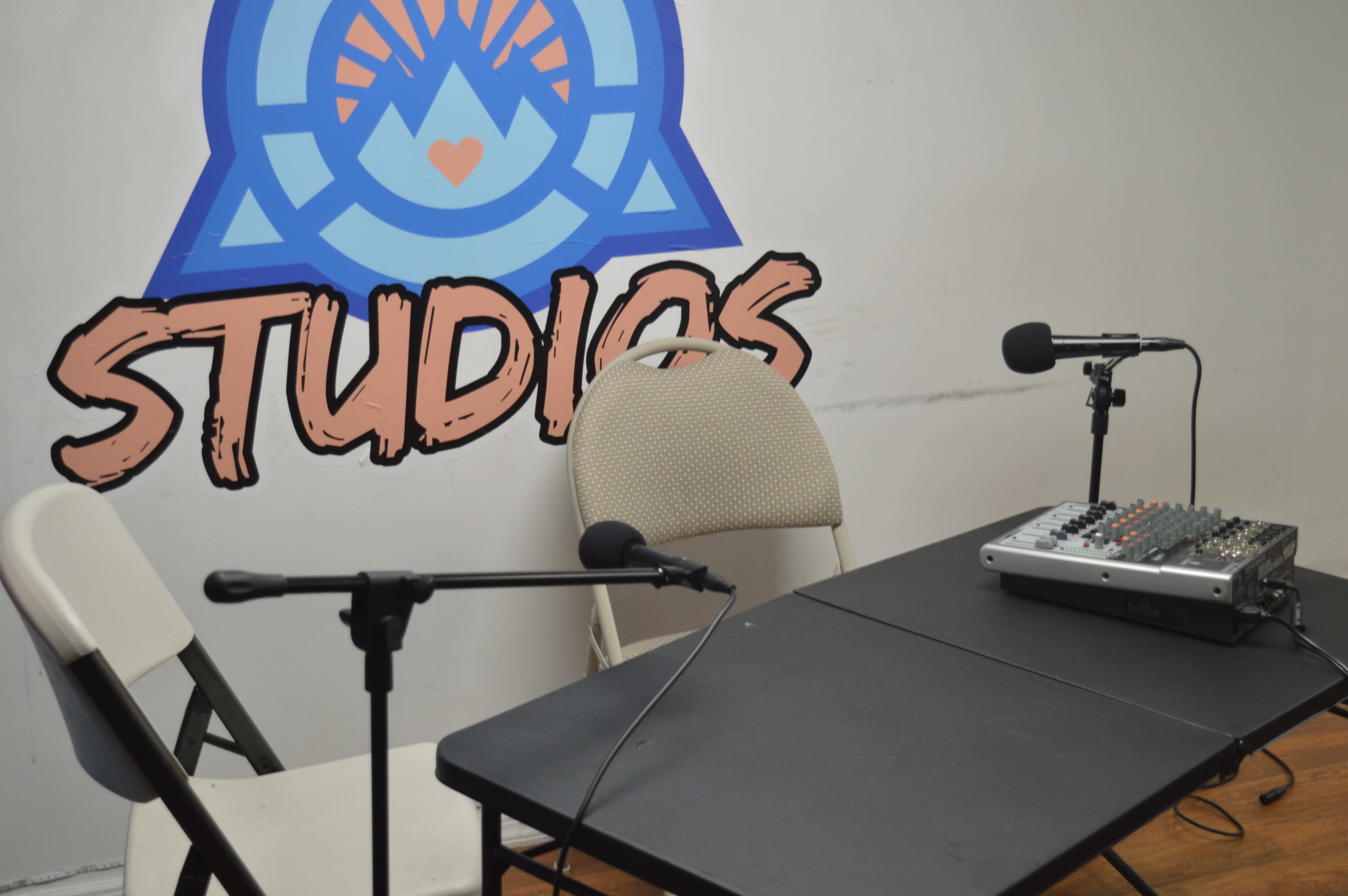 A small recording studio setup with two chairs, a small table, a sound mixer, and microphones, against a wall featuring a colorful stylized logo that says "STUDIOS."