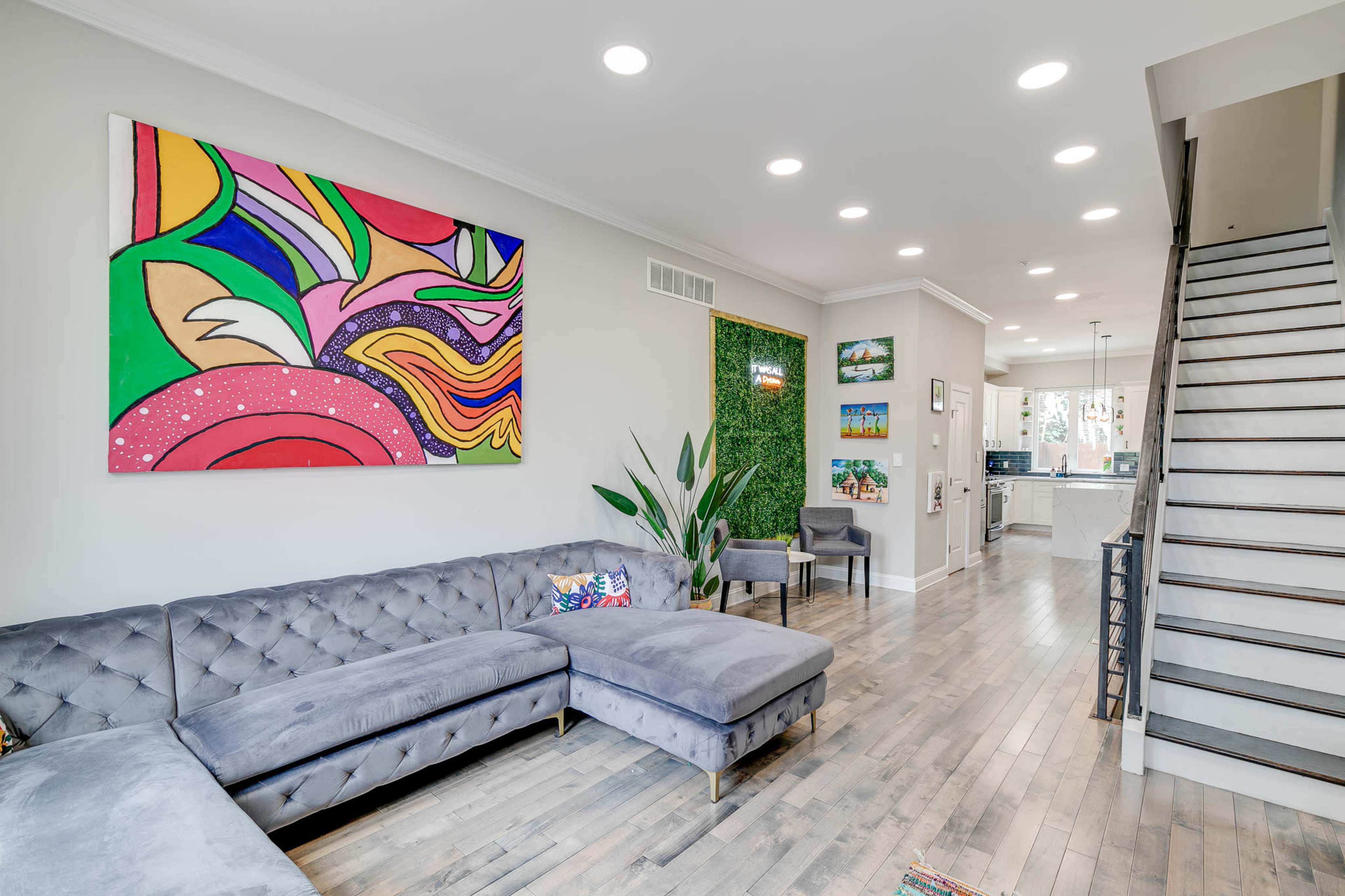 A modern living room with a gray sectional sofa, colorful wall art, a green accent wall, and a staircase leading to an upper level.