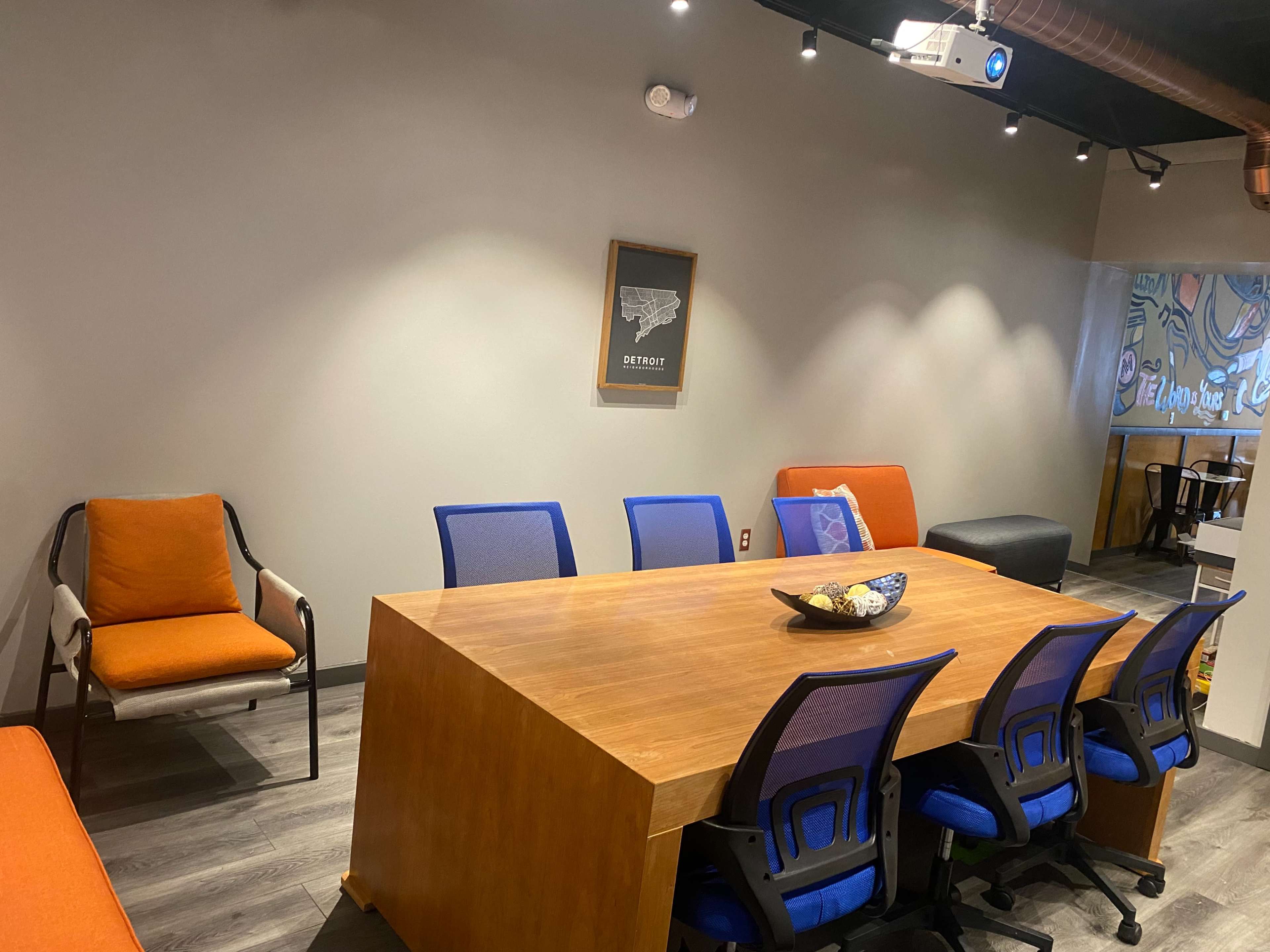 A modern conference room with a wooden table surrounded by blue chairs, an orange sofa in the corner, and a projector mounted on the ceiling.