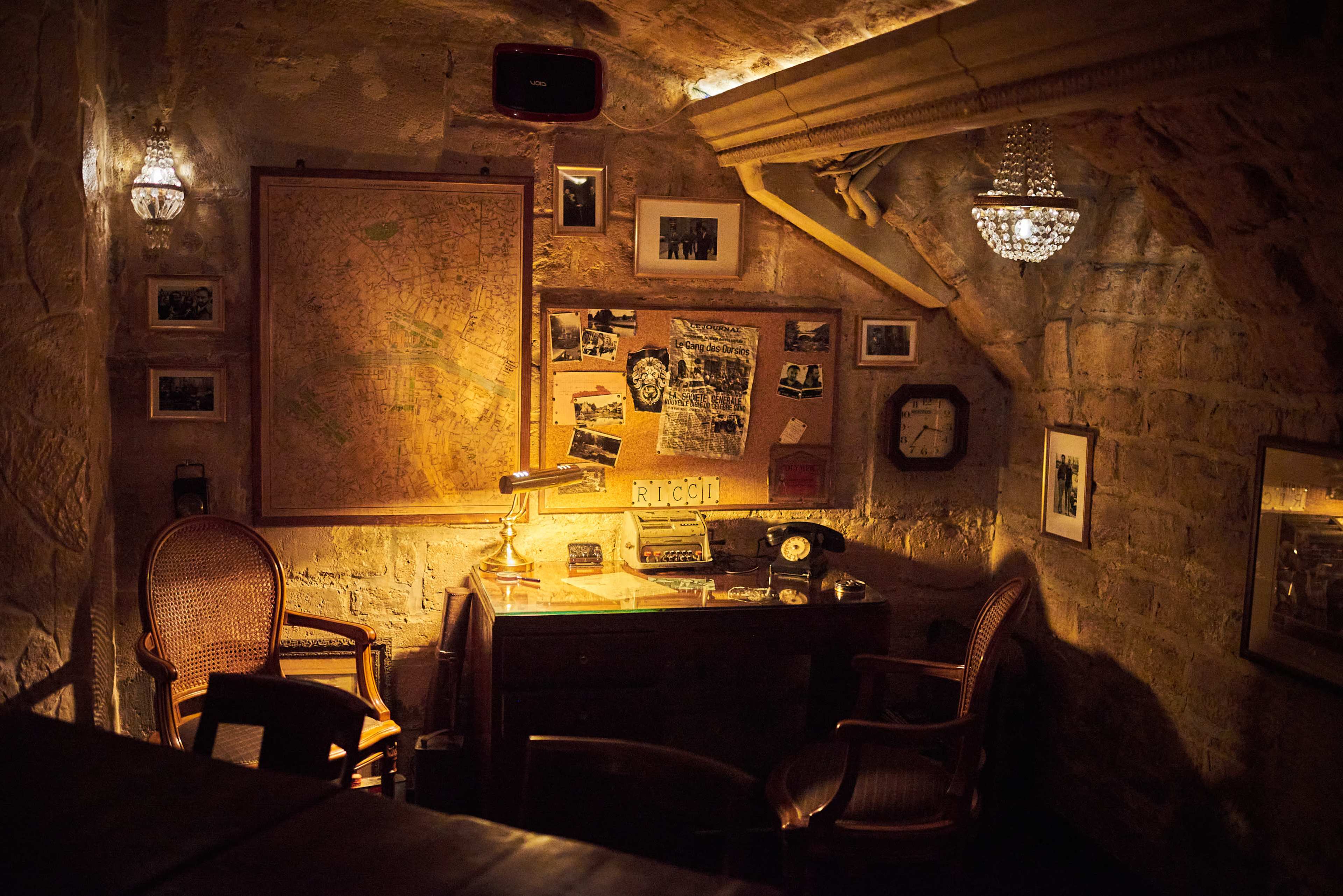 A dimly lit room features a wooden desk with a vintage lamp, old photographs, a rotary phone, and two wicker chairs against a stone wall adorned with a large map and framed images.