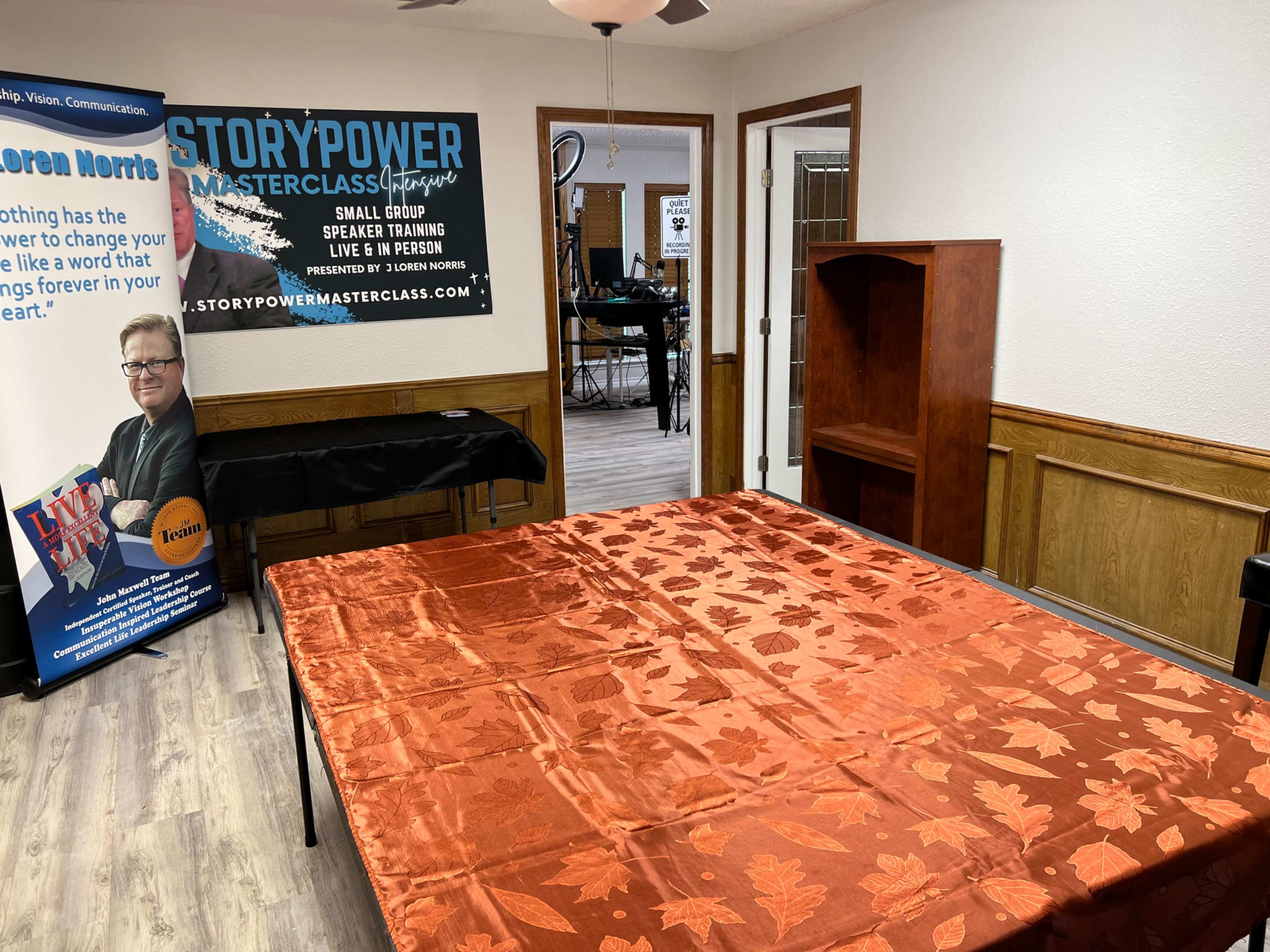 A training room with a large orange table covered in a leaf-patterned tablecloth, a banner featuring a speaker's photo, and a wooden shelf against the wall.