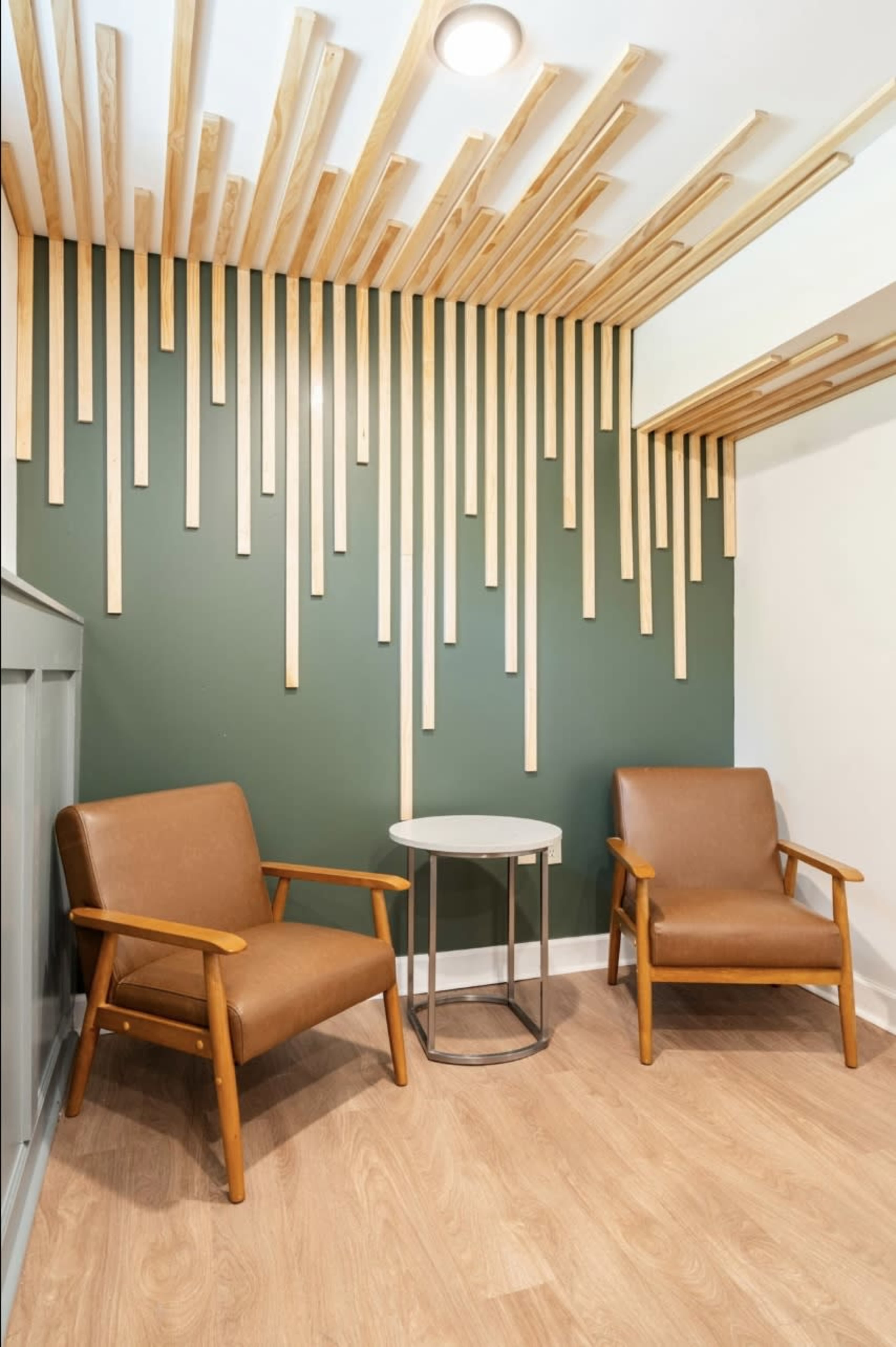 A small seating area with two brown armchairs and a round table, set against a green wall adorned with vertical wooden slats.
