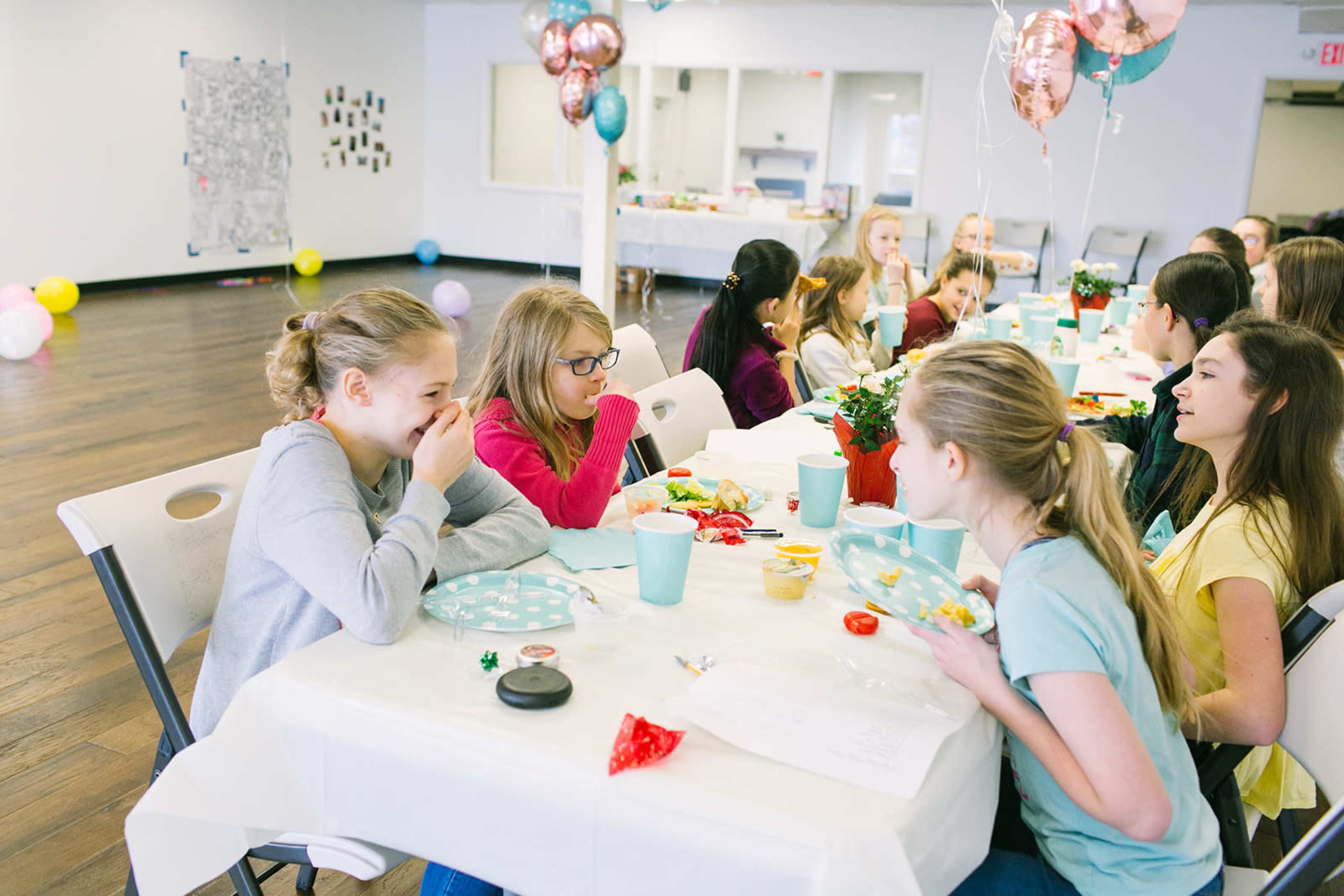 A group of young girls are seated at a long table enjoying snacks and conversation in a decorated room with balloons and a festive atmosphere.
