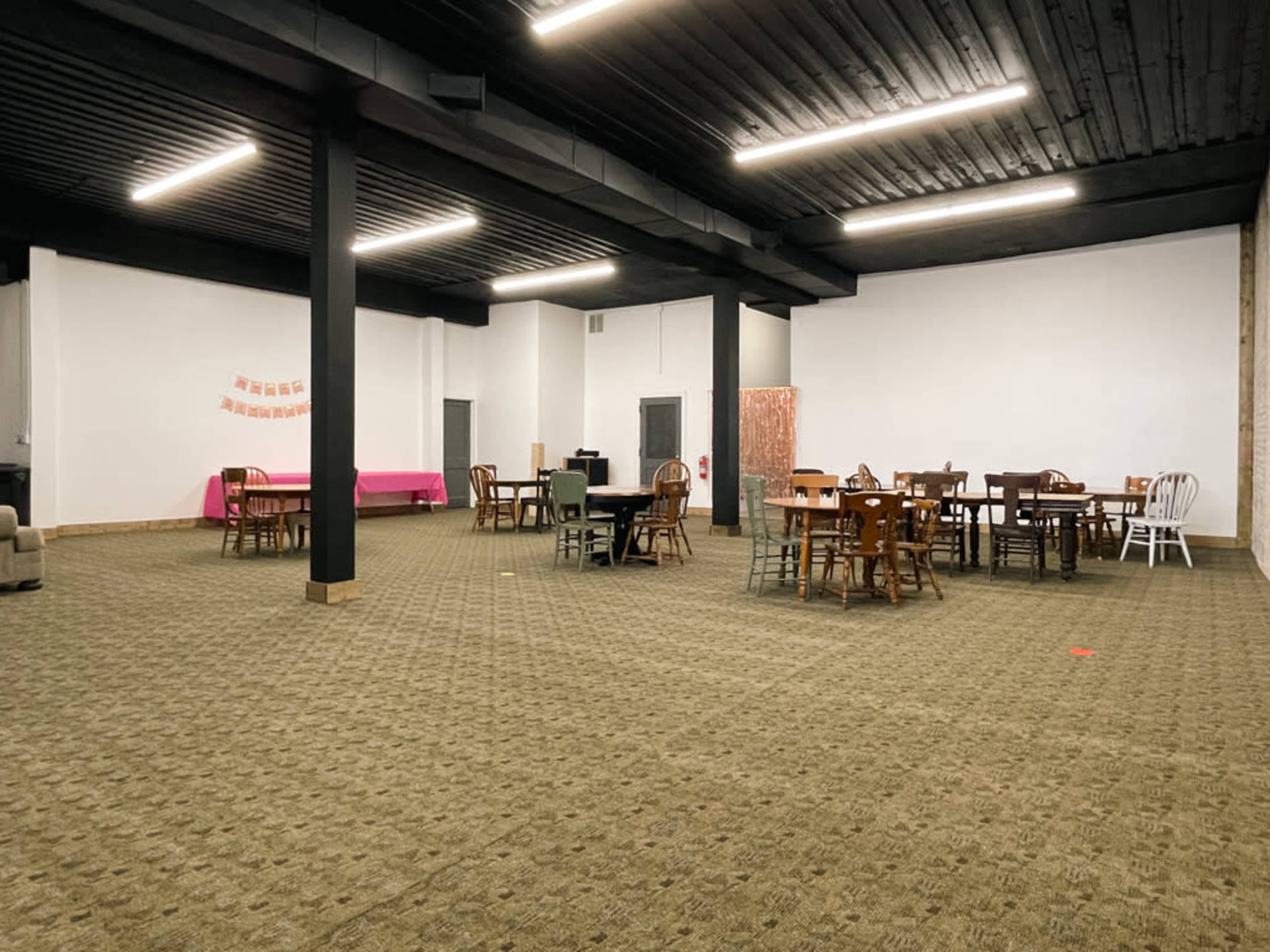 The image shows a spacious room with multiple wooden tables and chairs arranged throughout a carpeted floor, featuring black beams and white walls.