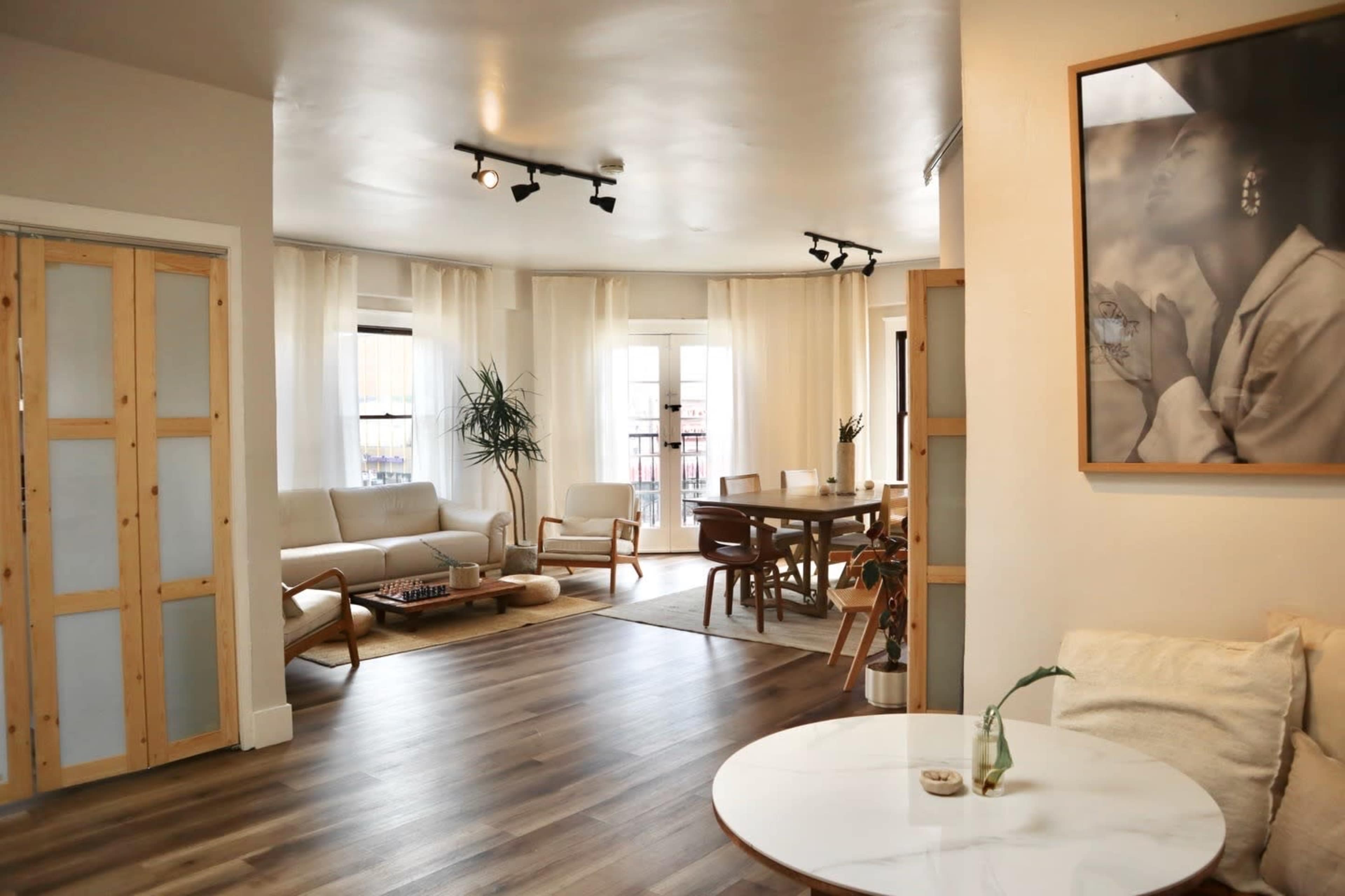 The image shows a spacious living area with light-colored walls, large windows, modern furniture, and a dining table set for several people.