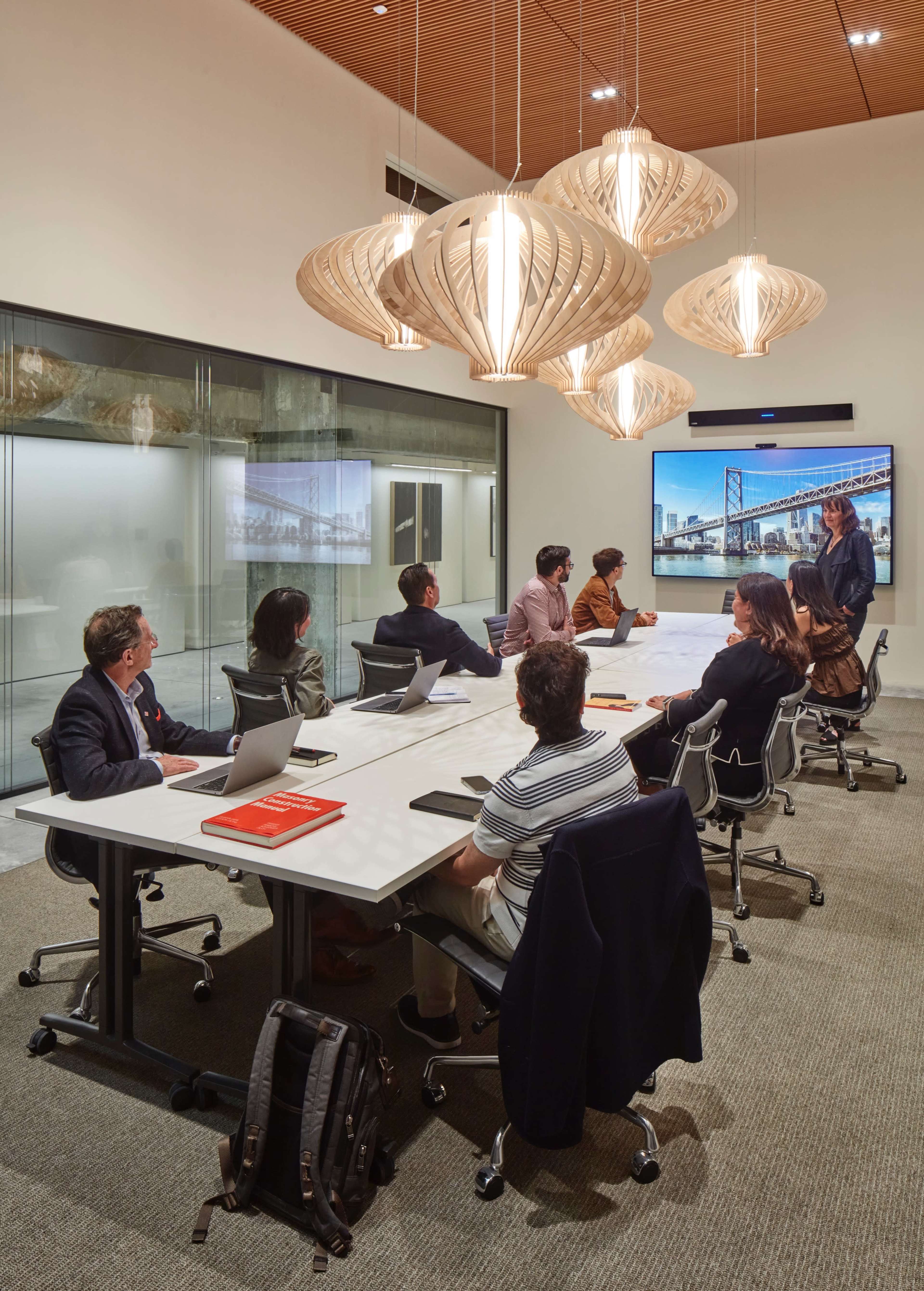 A group of eight people is seated around a large conference table in a modern meeting room, attentively observing a presentation on a screen.