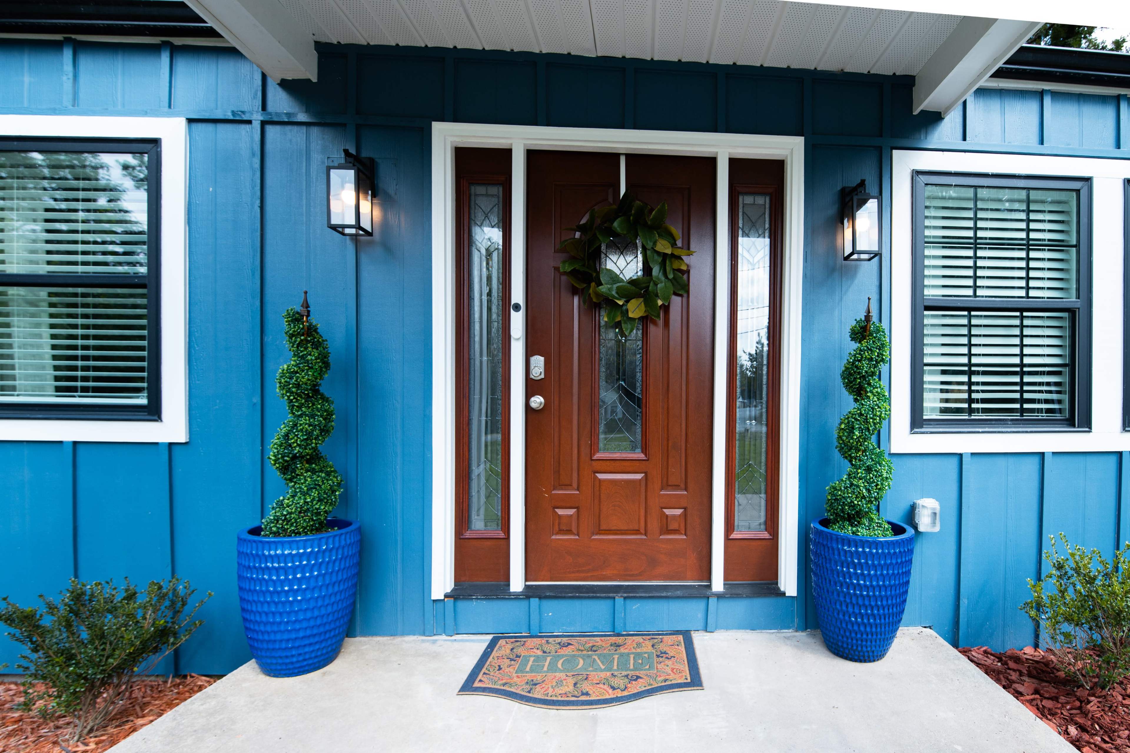 The entrance features a wooden double door flanked by two topiary plants in blue pots, with a welcome mat in front on a concrete walkway.