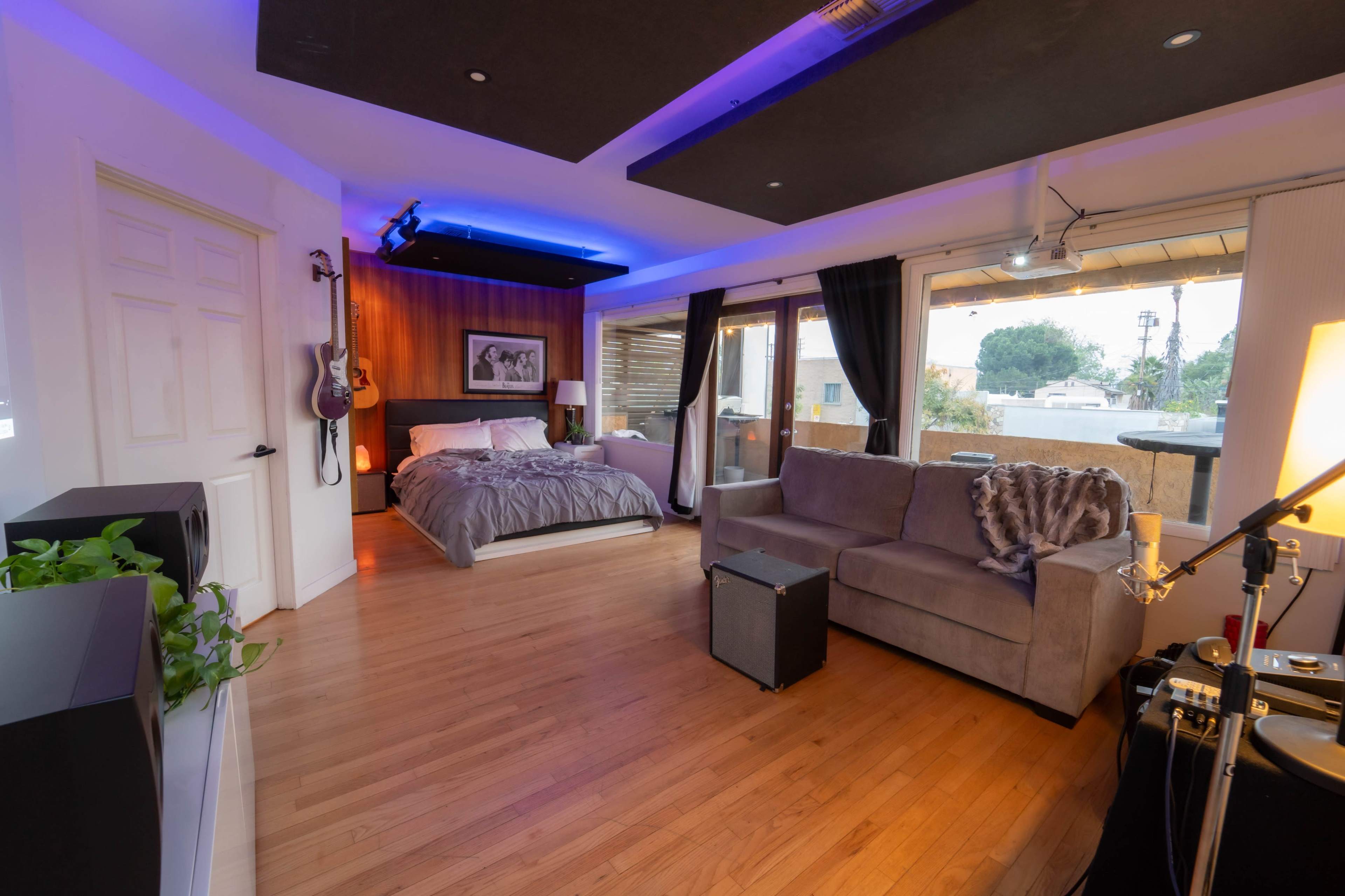 A modern studio apartment featuring a bed, a gray sofa, large windows, and a small audio setup.