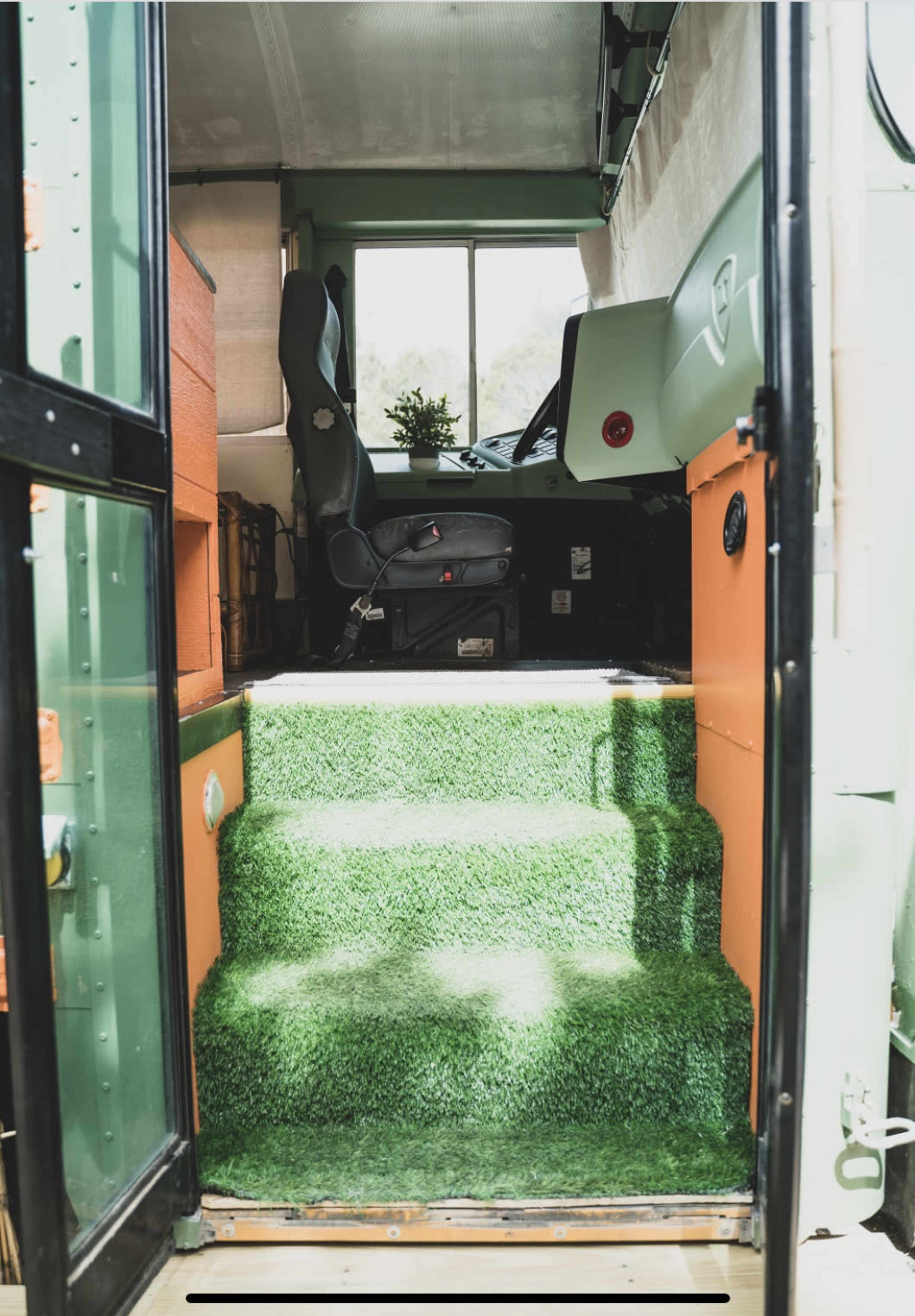 The interior of a converted bus with a grass-like carpet leading to a driver's seat near a large window.