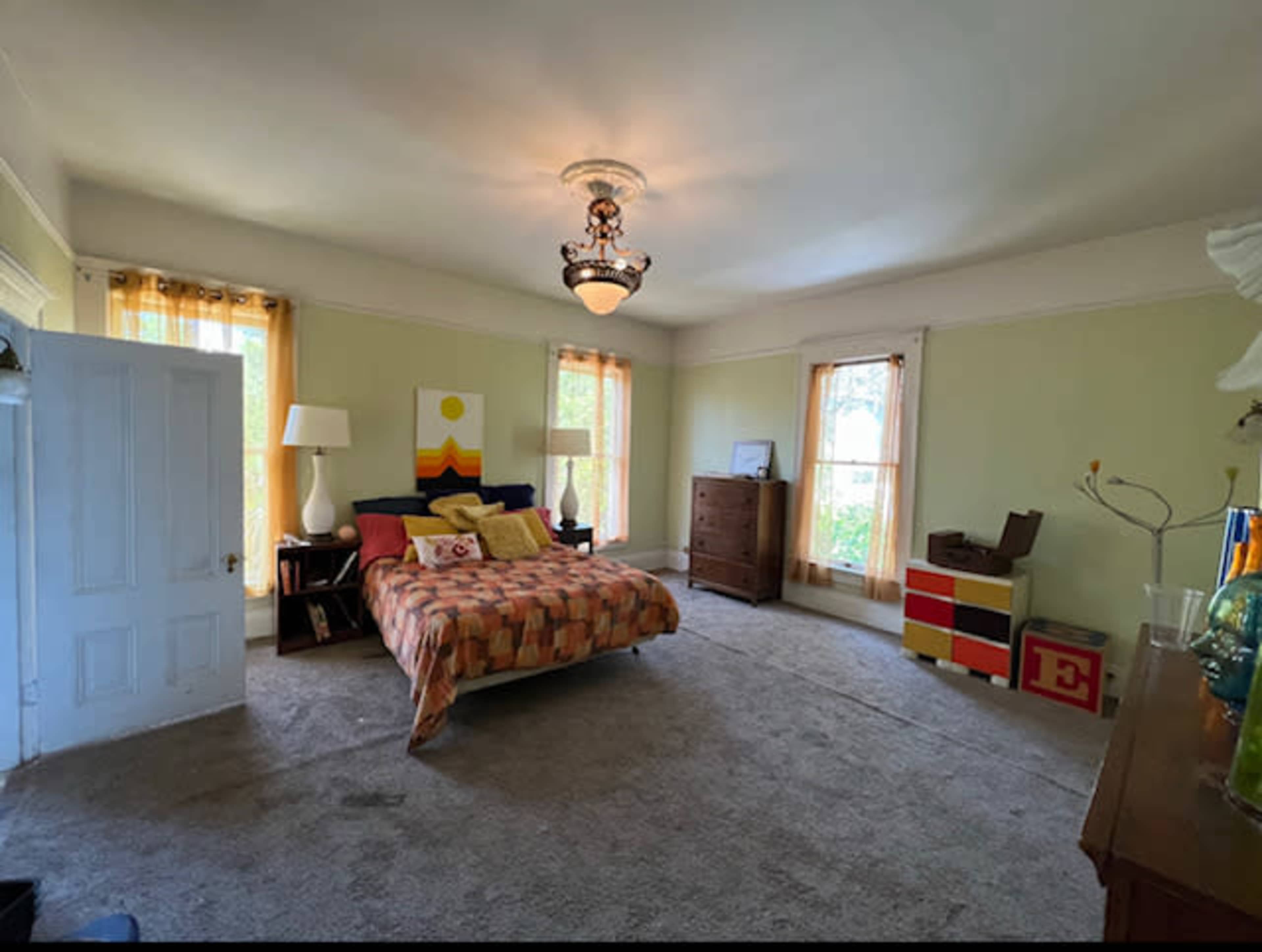 A spacious bedroom features a large bed with a patterned comforter, two lamps, a wooden dresser, and a colorful accent table against the walls.