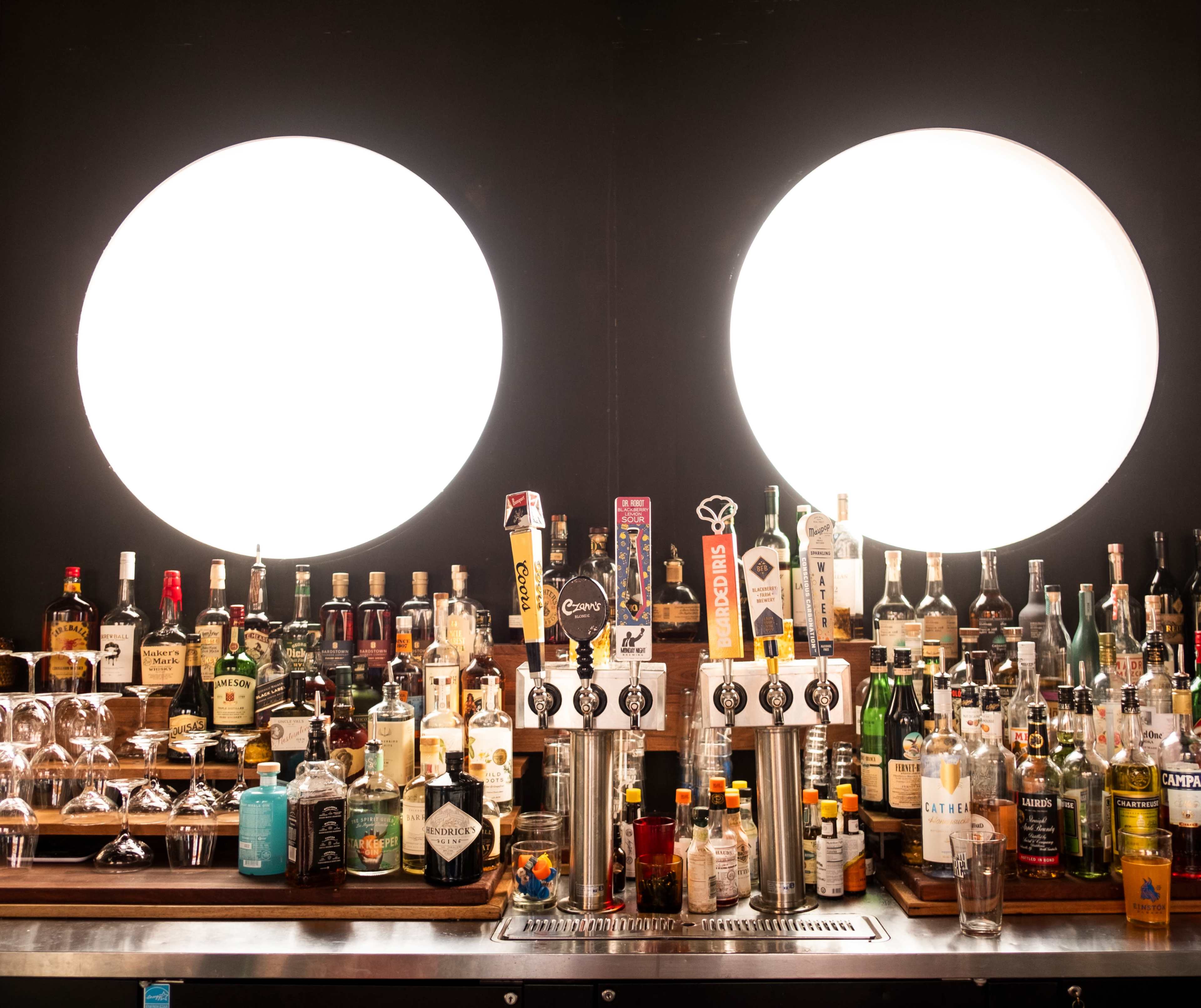 A bar counter is lined with various bottles of alcohol, with two large circular lights illuminating the scene.