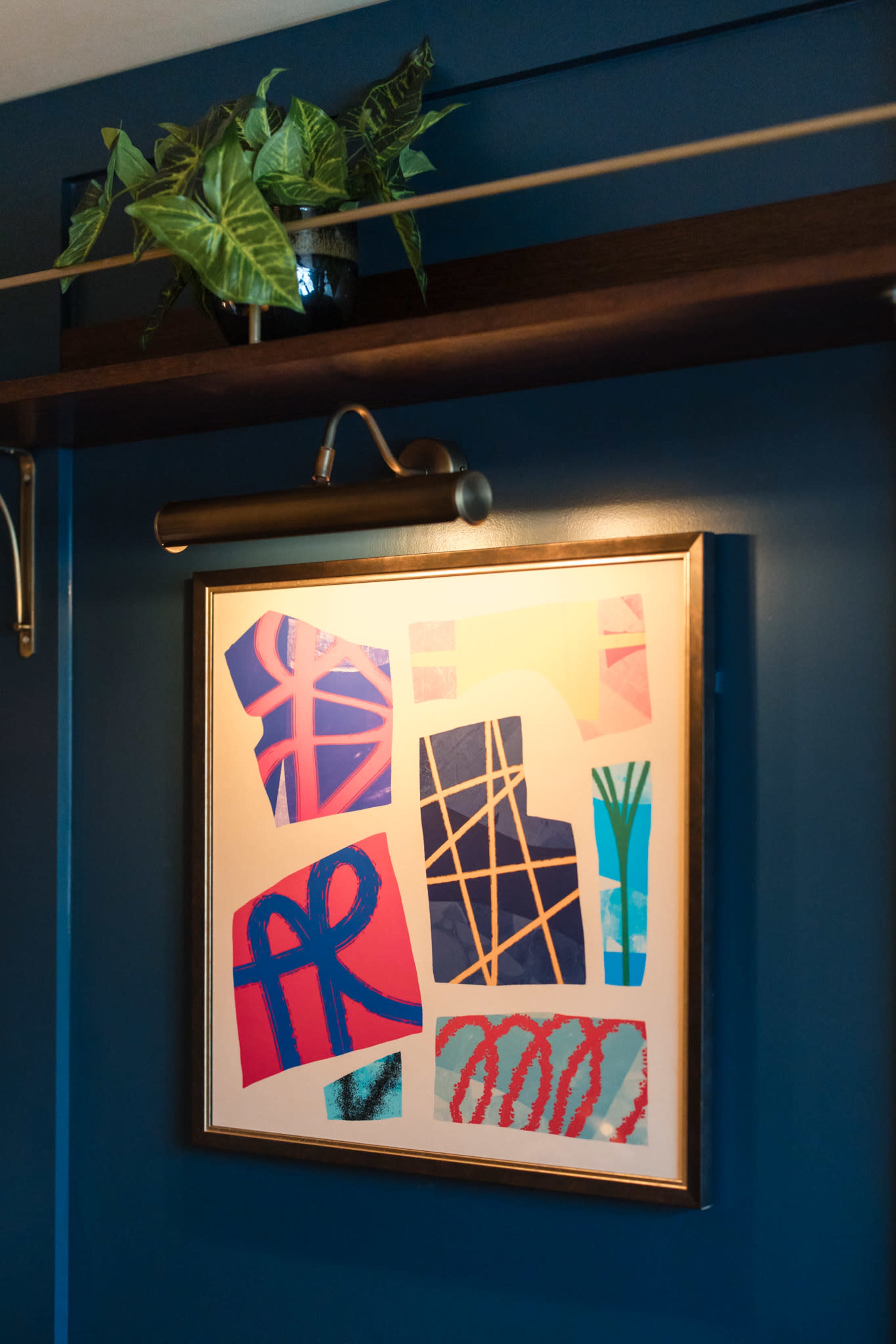 The image features a framed collage of abstract art displayed on a blue wall, with a potted plant positioned above it on a wooden shelf.