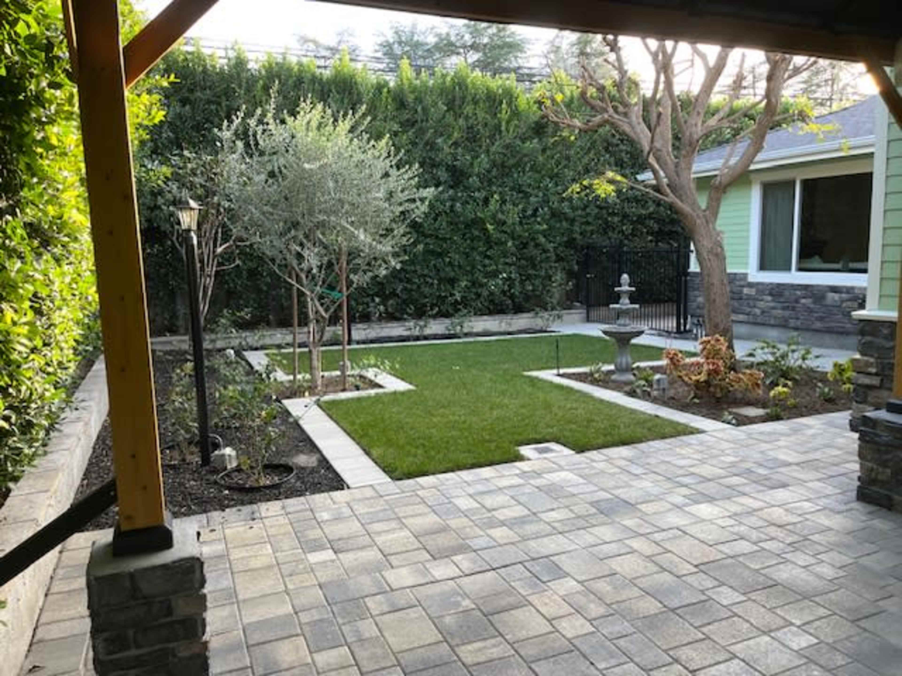 The image shows a landscaped backyard featuring a grassy area, trees, and a stone fountain, viewed from a covered patio.