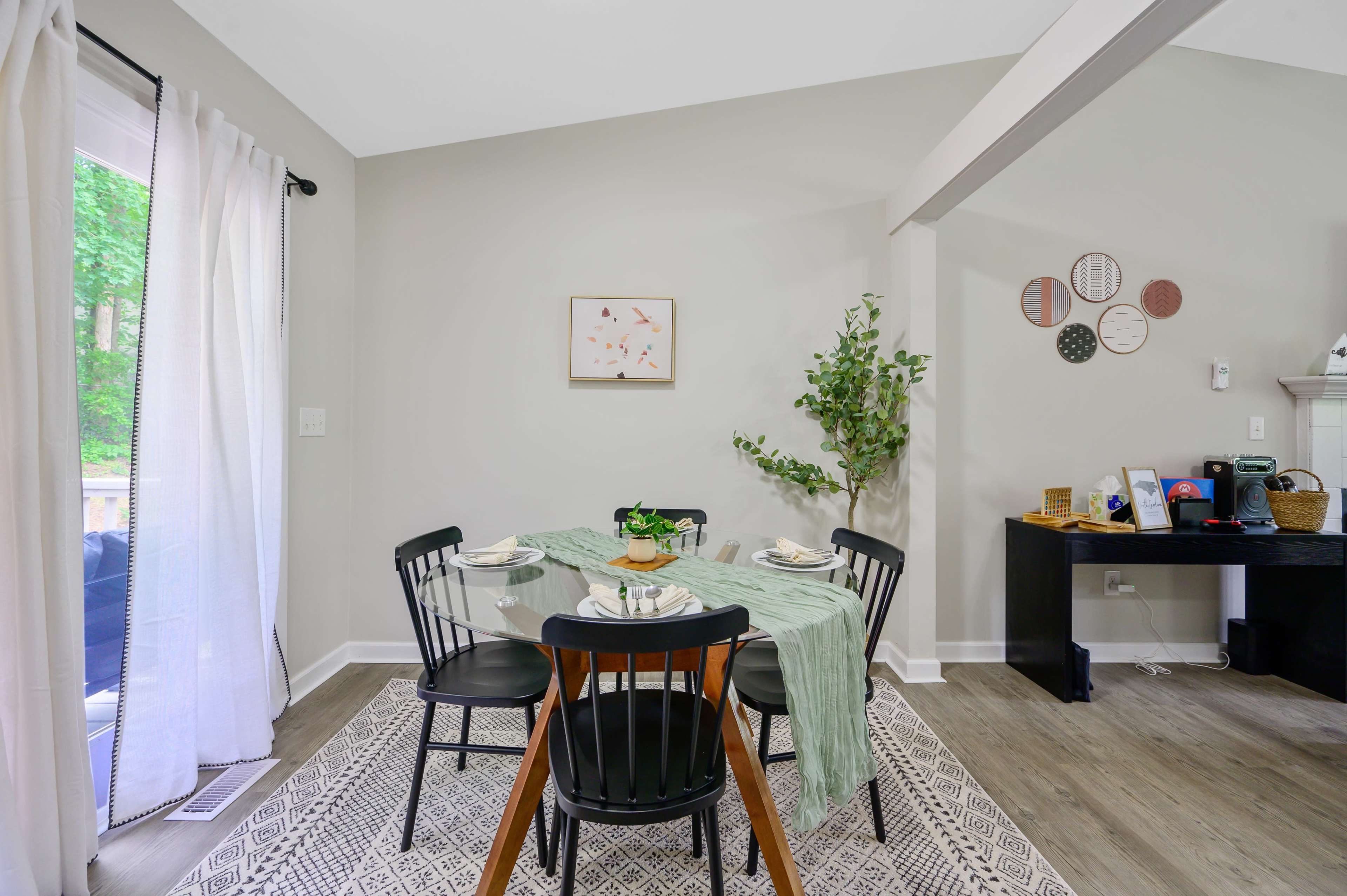 A dining area features a round table set for four, surrounded by black chairs, with a green table runner, plants, and decorative wall art.