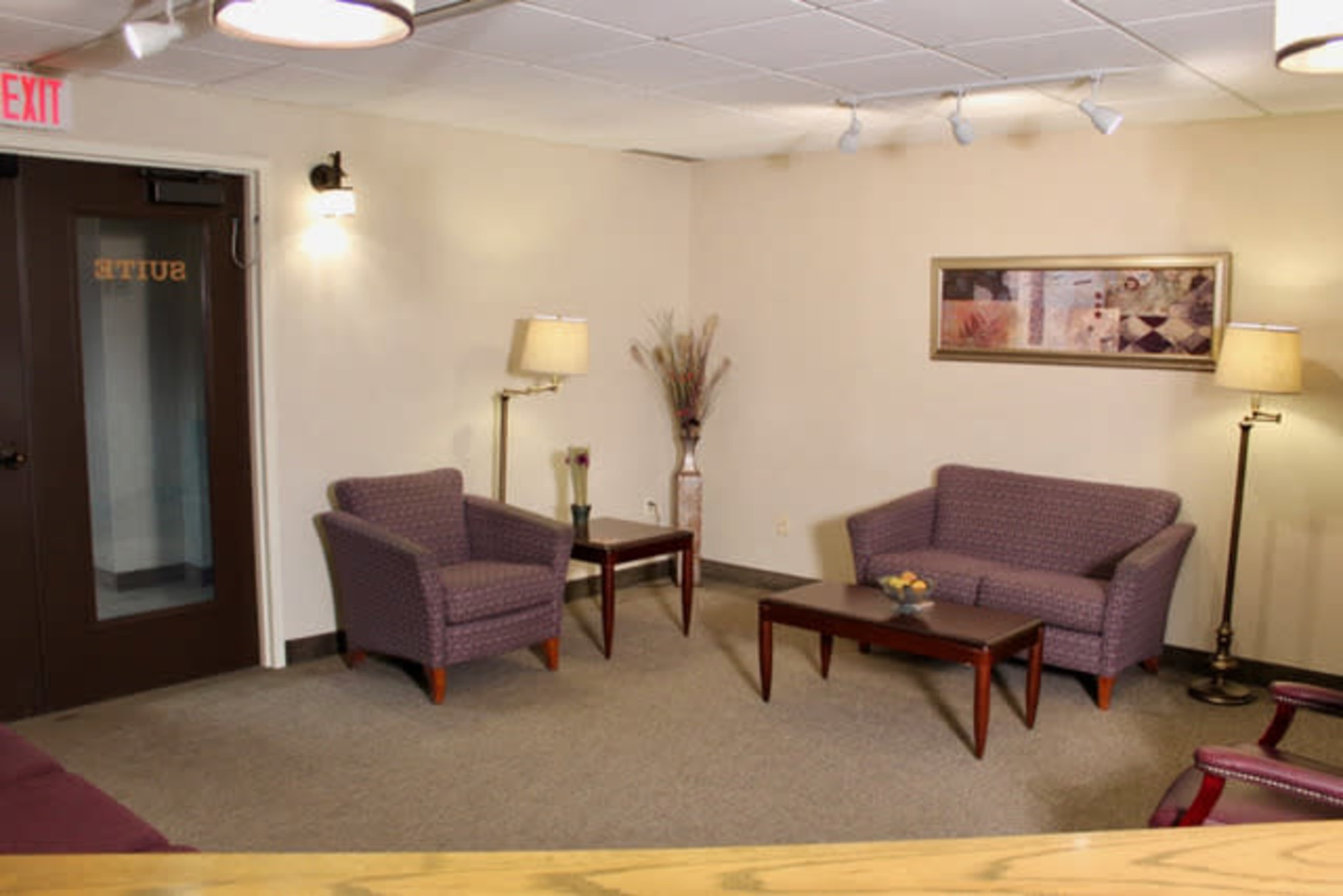 A waiting area with two purple armchairs, a brown coffee table, and lamps, accompanied by a decorative wall piece and a door labeled "SUITE."