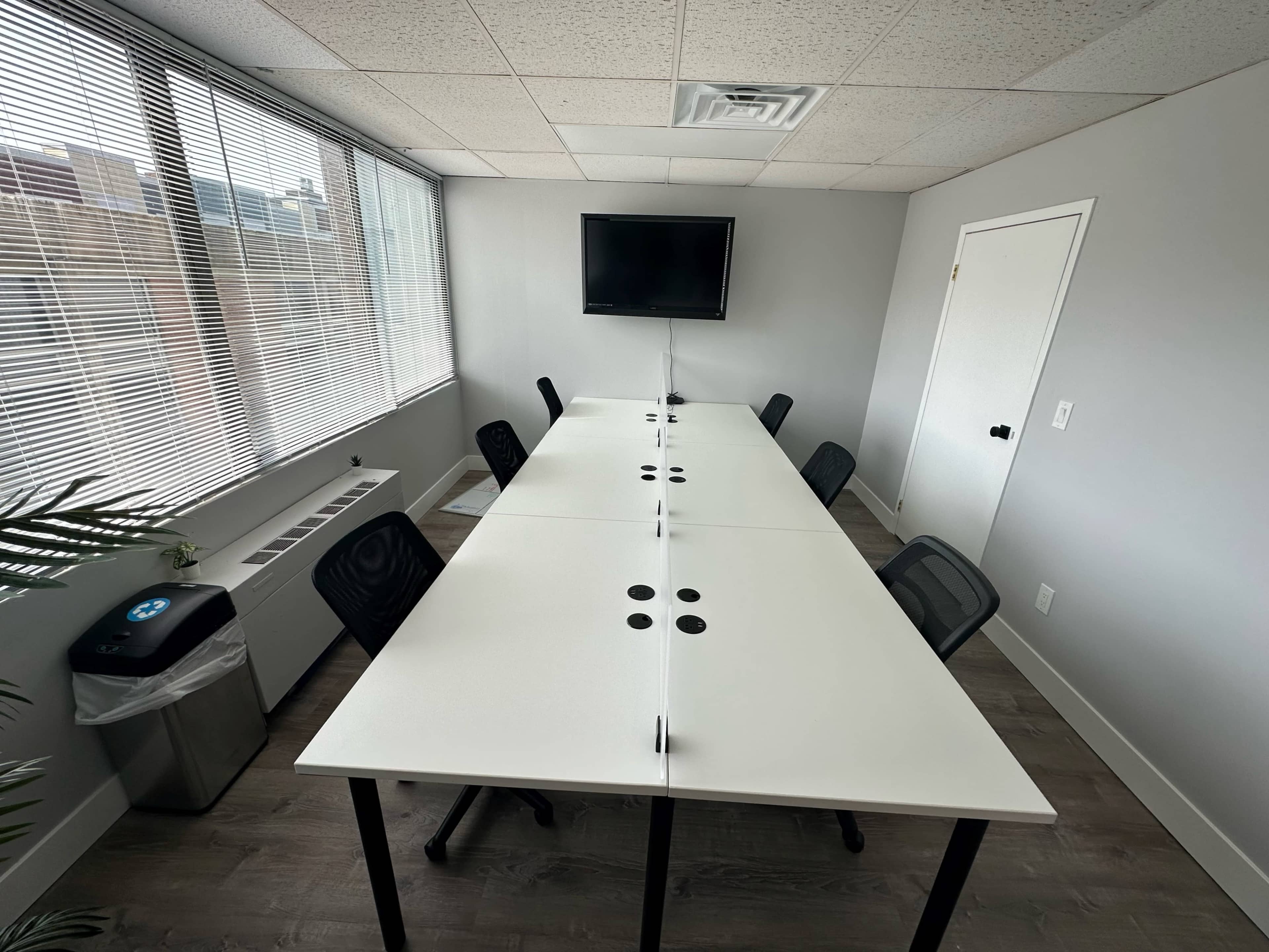 A long, white conference table with black chairs is positioned in a well-lit meeting room featuring large windows and a wall-mounted television.