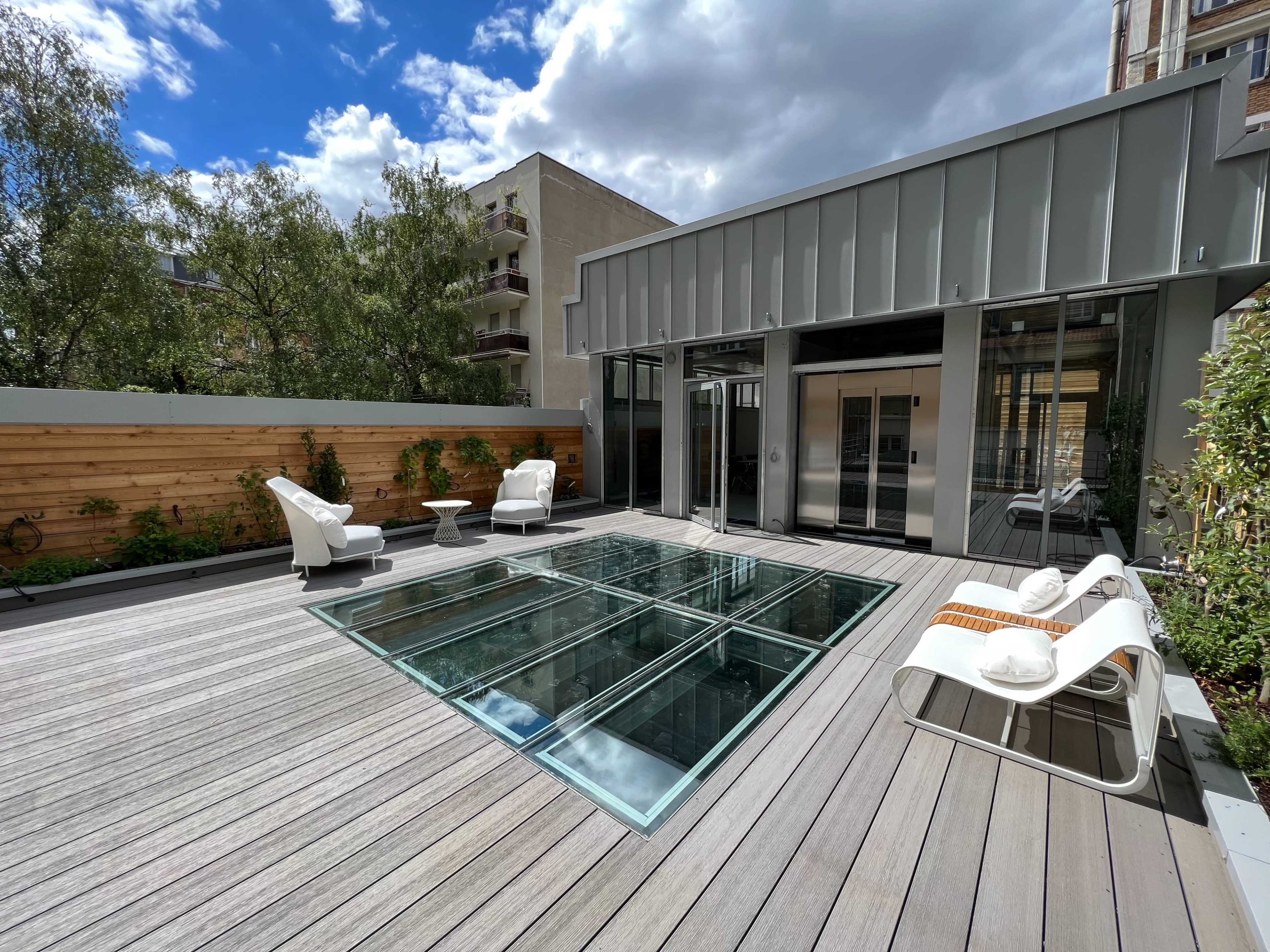 A modern rooftop terrace features a glass floor section, two white chairs, a small table, and surrounding greenery.
