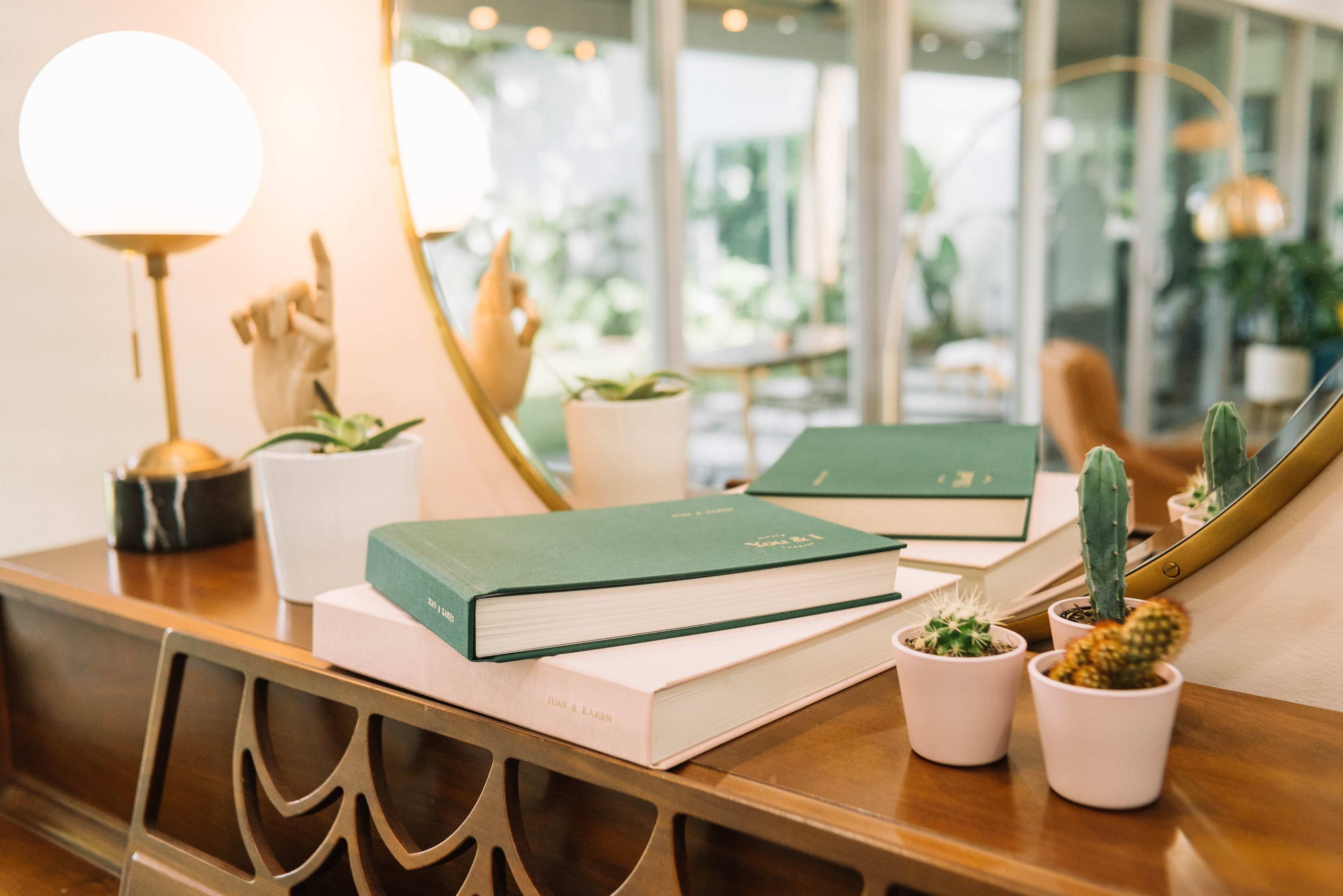 A wooden table displays stacked green books next to small potted cacti and a round mirror reflecting a bright, indoor space.
