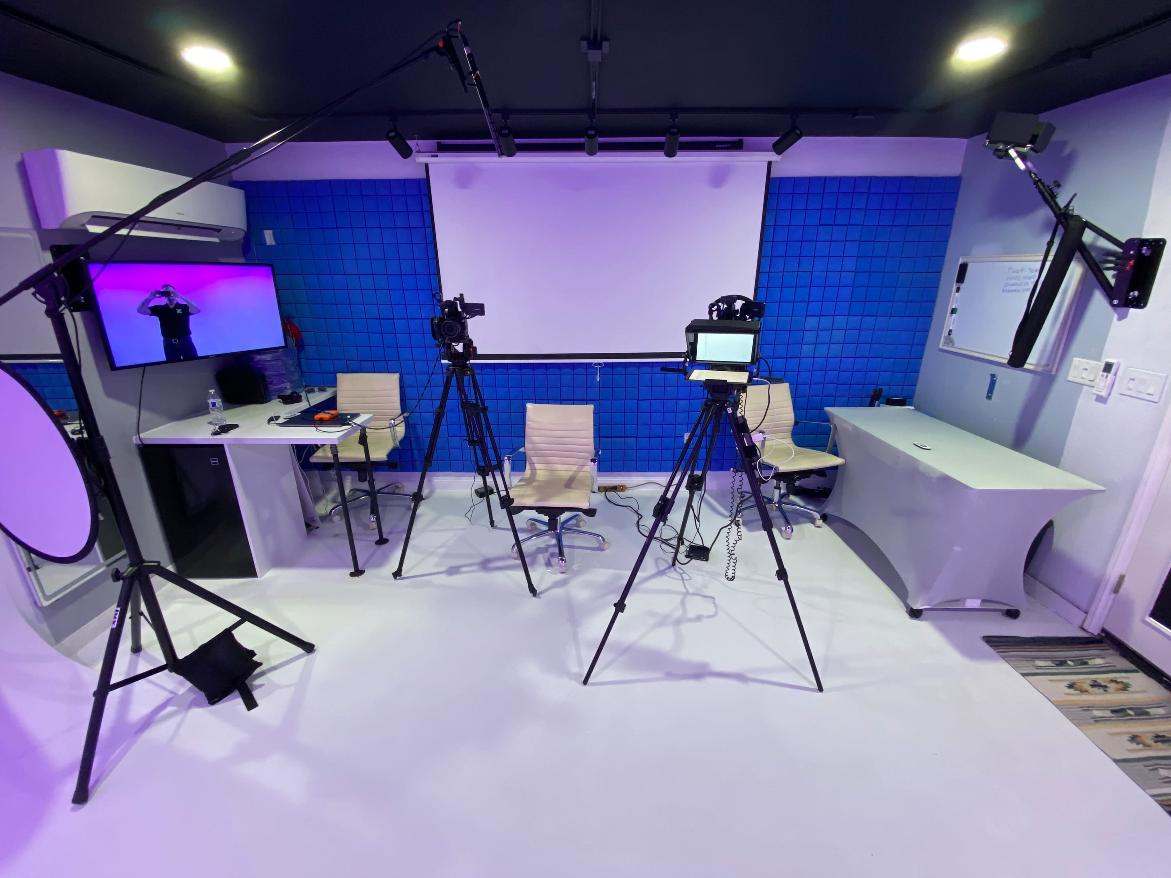 The image shows a studio setup with three camera tripods, two chairs, a white table, and a large screen against a blue wall.